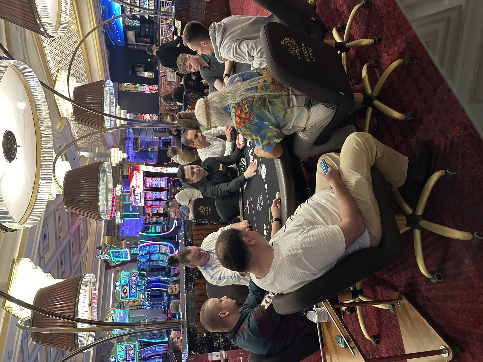 This image depicts a poker game taking place at a casino in Estosadok, Russia. The scene is set within a richly decorated gambling hall, characterized by ornate chandeliers and plush red carpeting. Several individuals are seated around a poker table, engaged in the game. The participants are a mix of men and one individual with long, white hair and a floral shirt. They are actively playing poker, with cards and chips visible on the table. In the background, slot machines and other casino attractions are apparent, suggesting a lively entertainment venue. The atmosphere appears focused on the game, with participants concentrated on their hands and the unfolding play.