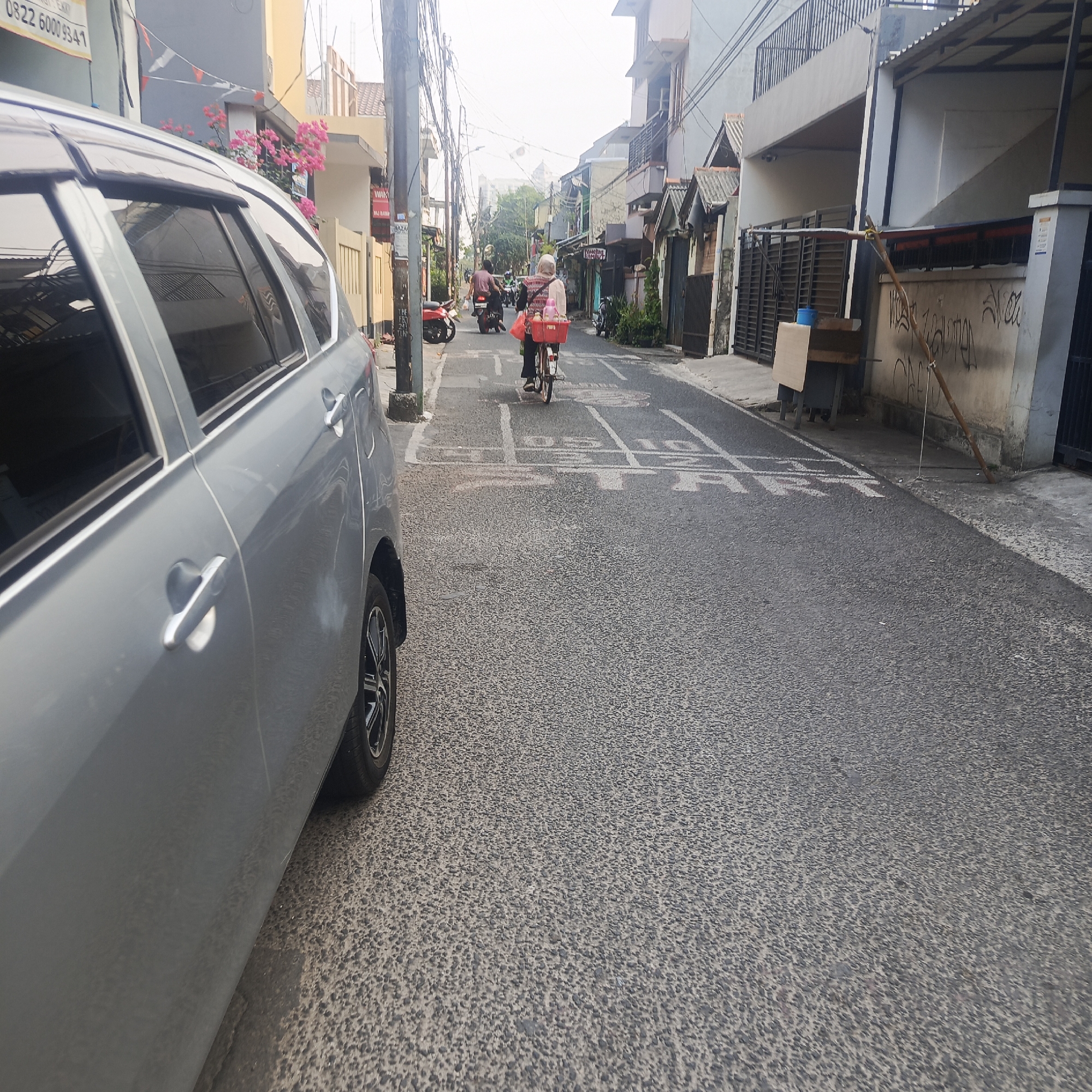 In South Jakarta, Indonesia, on a residential street, a silver car is parked on the left, facing away from the camera. A woman on a bicycle, wearing a headscarf and carrying a basket, cycles away from the camera down the middle of the narrow street. Several motorcycles are parked along the left side of the street. Houses with balconies and fences line both sides of the road, and electrical wires crisscross overhead. The street is paved with asphalt, and there are white lane markings painted on it. The sky is overcast.