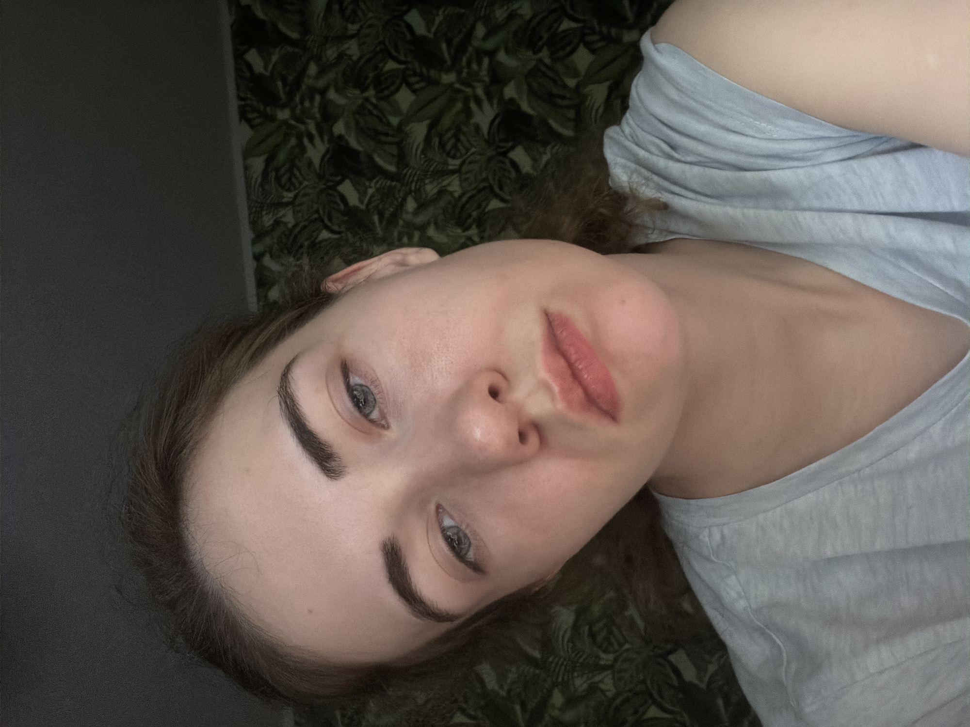 A close-up, frontal, medium shot captures a young woman with light skin and auburn hair against a dark, floral-patterned wallpaper. Her head is tilted slightly, and her eyes are looking downward with a somber expression. She is wearing a light blue t-shirt. The lighting suggests an indoor setting, possibly a bedroom, with a dark area in the upper left corner and the wallpaper filling the background on the right. The scene appears to be in Zavodoukovsk, Russia, likely within a residential dwelling.