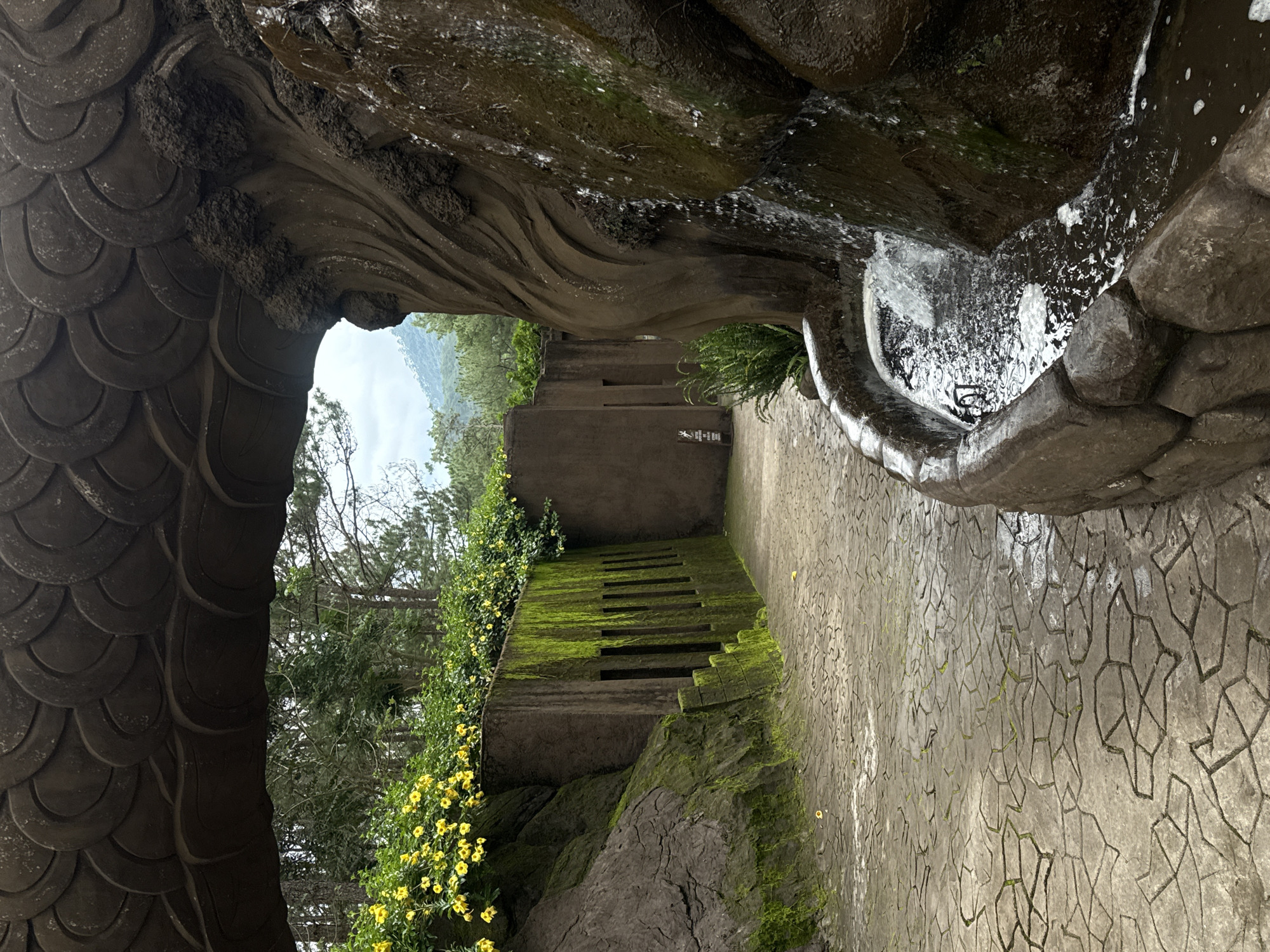 The image shows a pathway and a fountain in what appears to be a garden or park setting. The pathway is paved with irregularly shaped stones, some of which are wet. To the left of the path, a rock face covered in green moss slopes upwards, topped by a wall with several rectangular openings and a cluster of yellow flowers. Overhead, a textured, dark structure with a scale-like pattern arches across the scene. To the right, a fountain cascades down a rough, dark brown rock formation into a stone basin filled with water. In the background, trees and hills are visible under a cloudy sky. The overall impression is one of a decorative outdoor space, possibly a tourist attraction, given the artificial rockwork and structured plantings. While the exact location in Da Lat, Vietnam is not provided, such features are common in the city's gardens and attractions. There are no people or animals visible in the image.