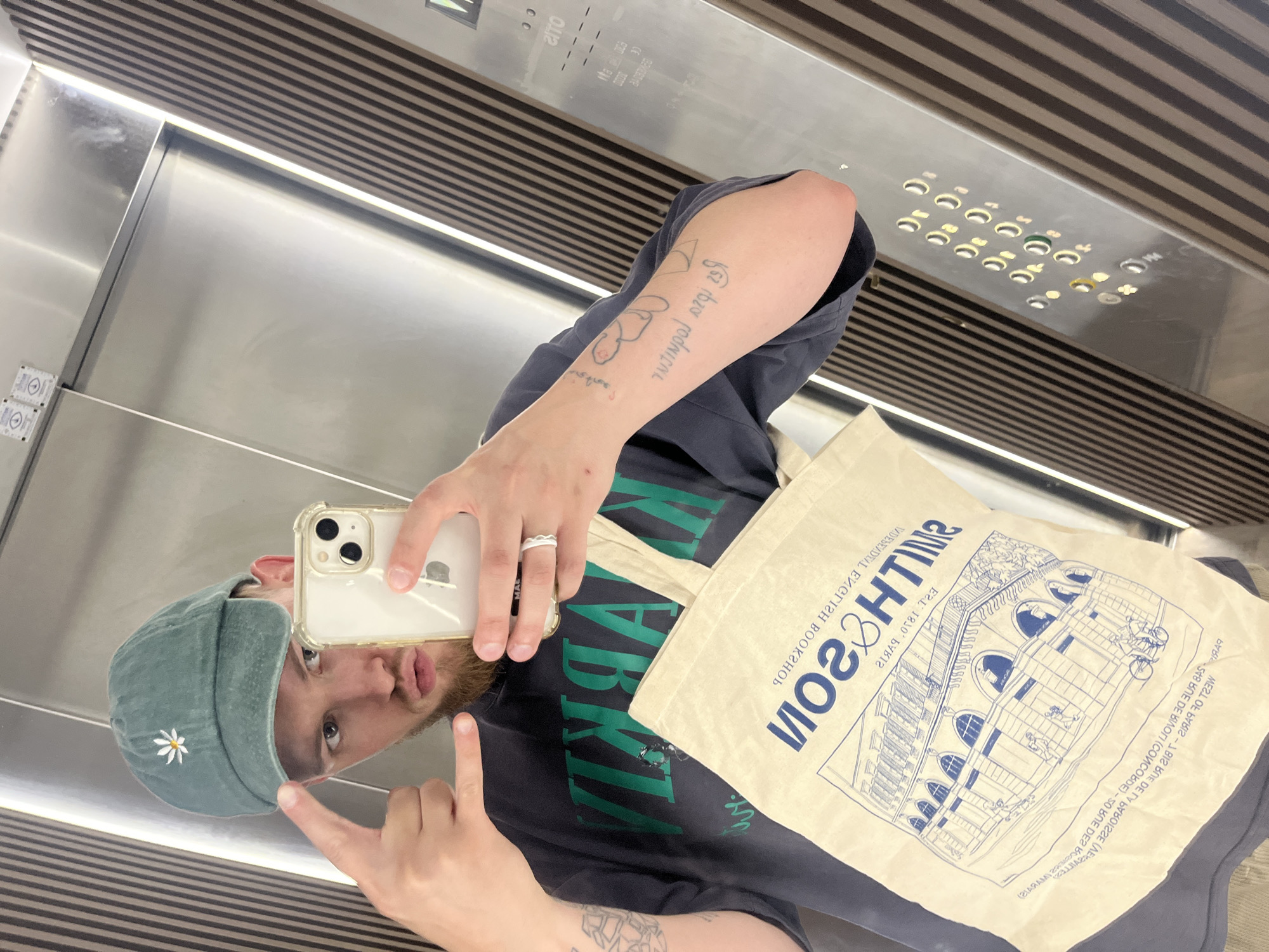 A man is taking a mirror selfie in what appears to be an elevator. He is wearing a dark t-shirt with green lettering and a light-colored tote bag with a blue drawing of a building and text is draped over his shoulder. He has on a teal baseball cap with a white daisy embroidered on the front. His right arm is visible and shows several tattoos. He is holding a smartphone in his left hand, taking the picture in a mirror. The elevator has stainless steel walls and a slatted ceiling. The text on the tote bag is in English and French, reading "SMITHSON ENGLISH BOOKSHOP" and referencing Rue de Rivoli and Concorde in Paris. The man has a beard and is looking at the camera with a playful expression, one hand giving a rock-on sign. The lighting suggests it is daytime.