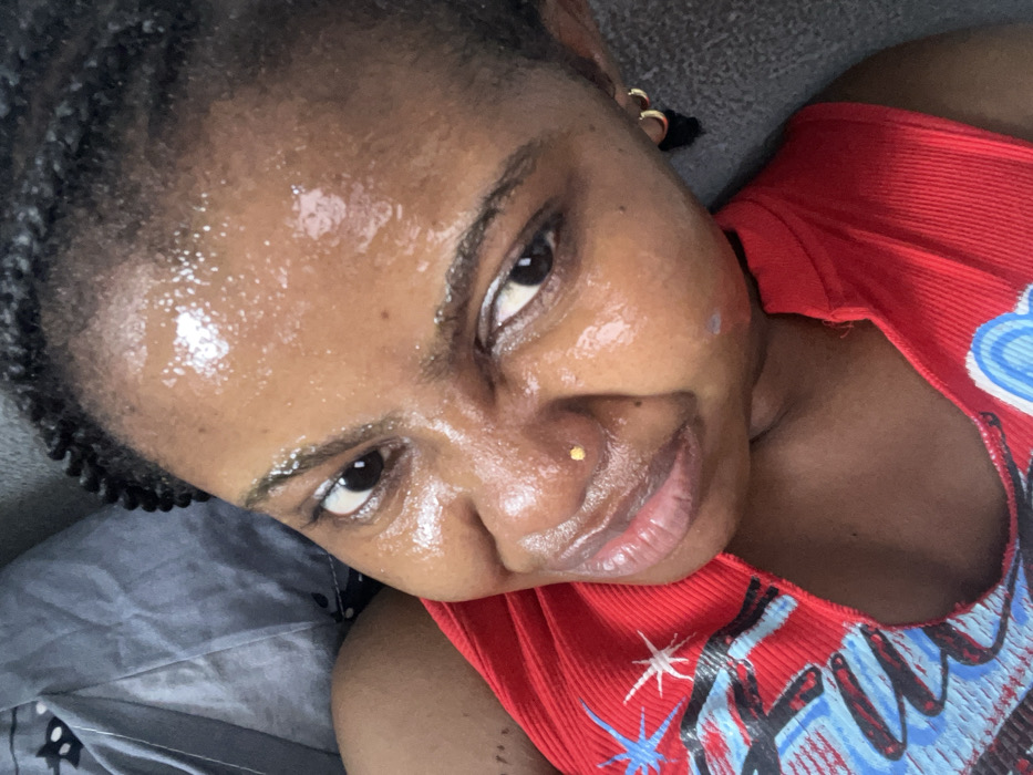 A female individual is depicted in a close-up, high-angle shot within the city of Ipetumodu, Nigeria. Her dark skin is noticeably moist or oily across her forehead, nose, and cheeks, reflecting ambient light. Her dark hair is styled in multiple small braids. A small, gold-colored stud pierces her left nostril. Small, gold-colored hoop earrings are visible on her right earlobe. She wears a red, ribbed sleeveless top with a blue and white patterned design on the left side of the chest. Her eyes are dark and focused directly forward, with a slight, closed-mouth smile on her lips. A faint, light-colored mark is present on her left cheekbone. The background consists of a crumpled grey fabric, possibly bedding, to the left, and a darker, textured grey surface to the right.
