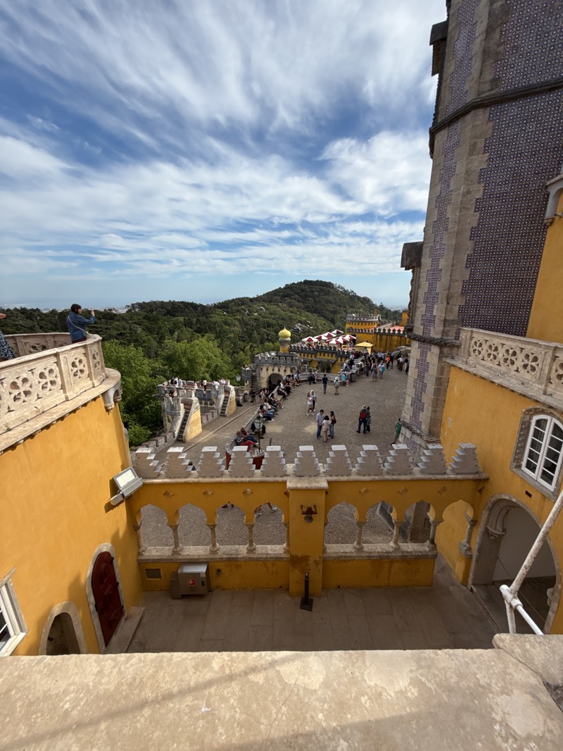 The image captures a view of Pena Palace in Sintra, Portugal. The perspective is from a high vantage point, looking down into a courtyard within the palace complex. The courtyard is filled with people, some walking around and others seated along the edges. The architectural features are predominantly yellow, with arched walkways and decorative crenellations. A tower with blue and white tiled accents is visible on the right side. Beyond the courtyard, there is a landscape of green trees and hills, with a cloudy sky above.