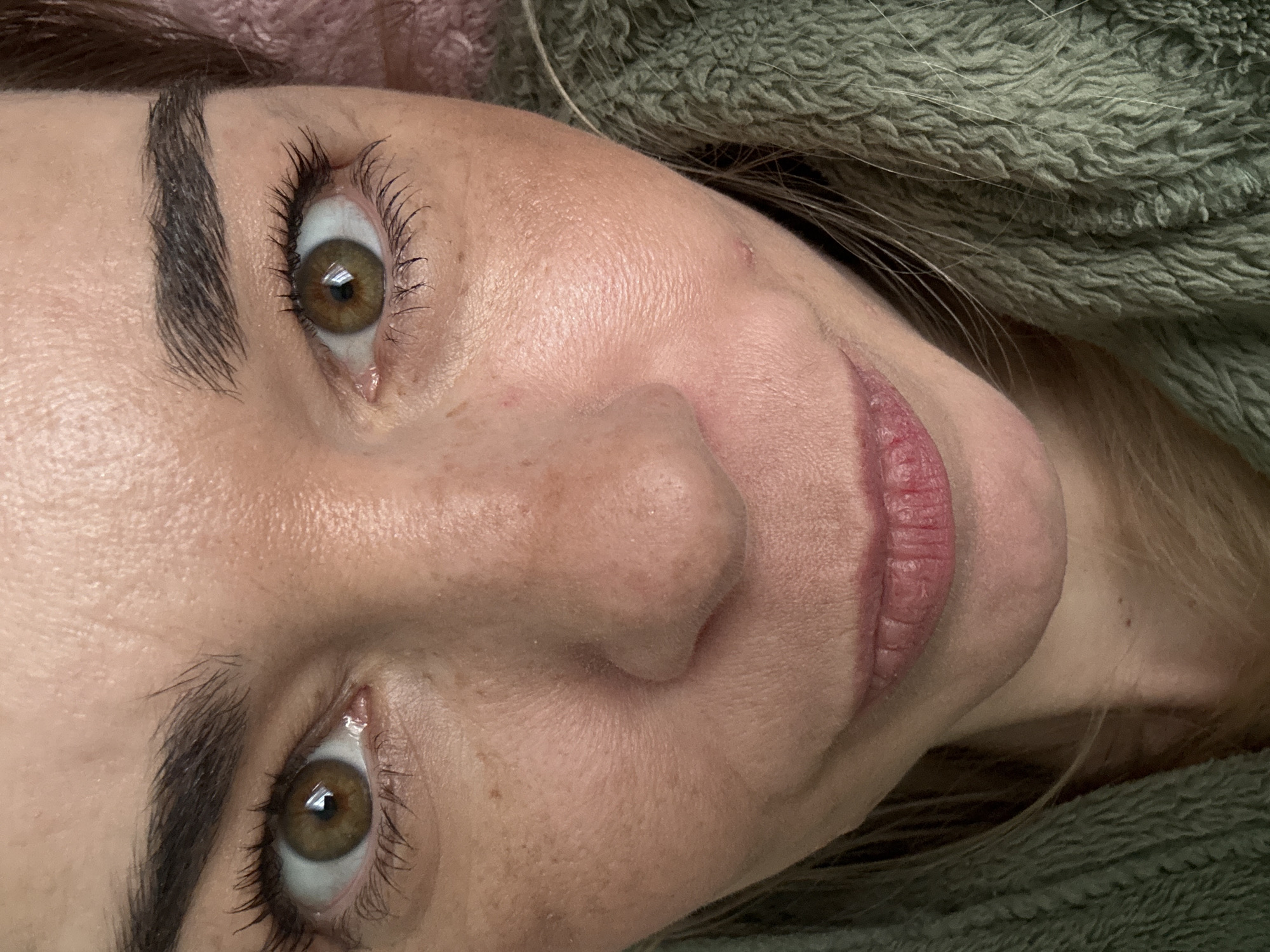 The image shows a close-up of a woman's face. She has fair skin, brown eyes, and dark eyebrows. Her lips are pink and appear slightly chapped. There are visible eyelashes with mascara applied. A few freckles are scattered on her nose and cheek. The background consists of soft, textured fabrics in shades of green and pink. There is no indication of the location being Vsevolozhsk, Russia, or any other specific place.
