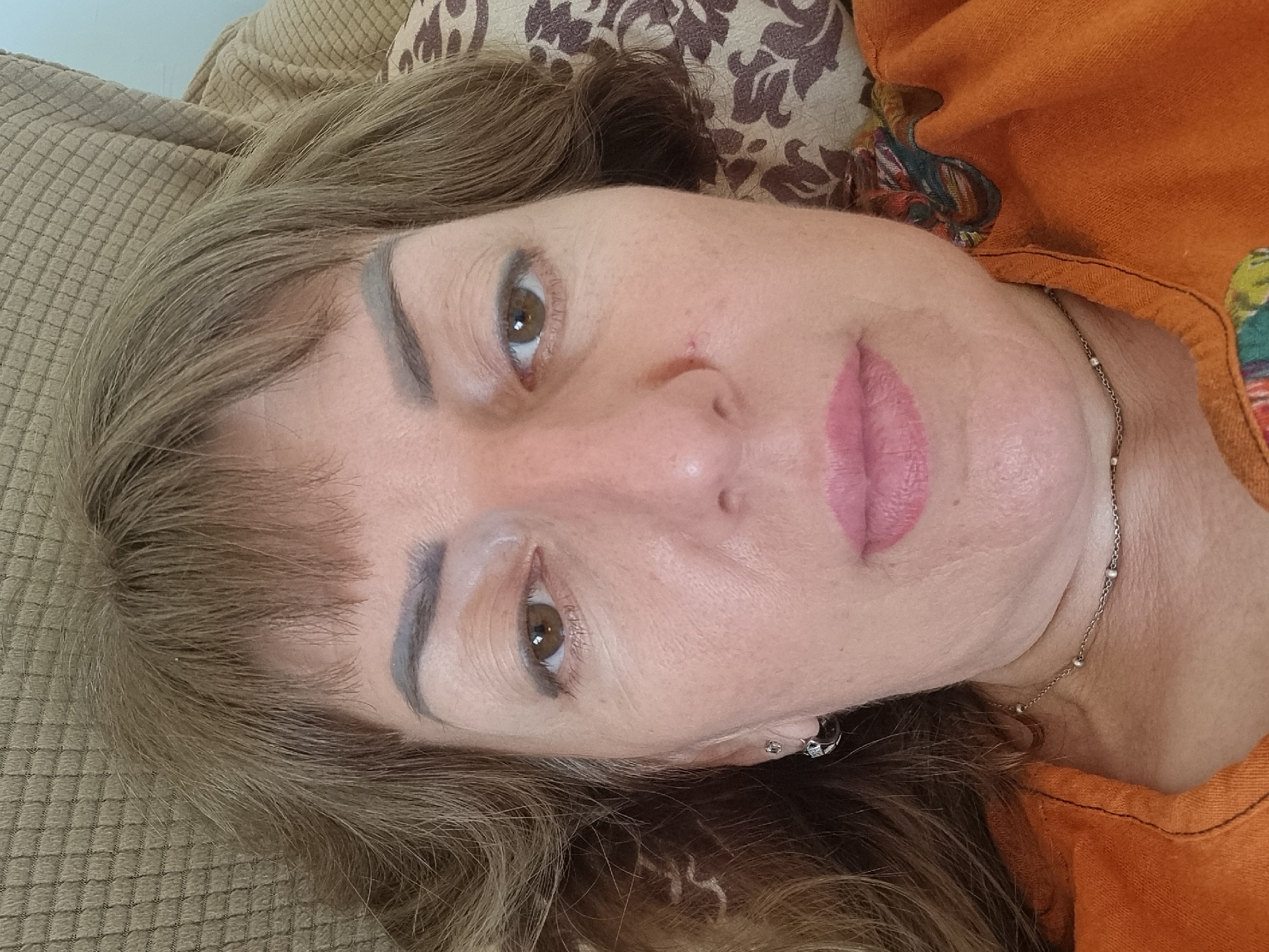 A selfie shows a Caucasian woman in her late 40s or 50s with medium-length brown hair and bangs. She is lying down, with her head resting on a brown textured surface, possibly a sofa or chair. The woman is wearing an orange shirt with a patterned neckline and a delicate silver necklace. Her left ear is adorned with a silver hoop earring. She appears to be indoors, with a patterned pillow visible behind her head. The overall impression is a casual, unposed moment. The location is not specified in the image itself.