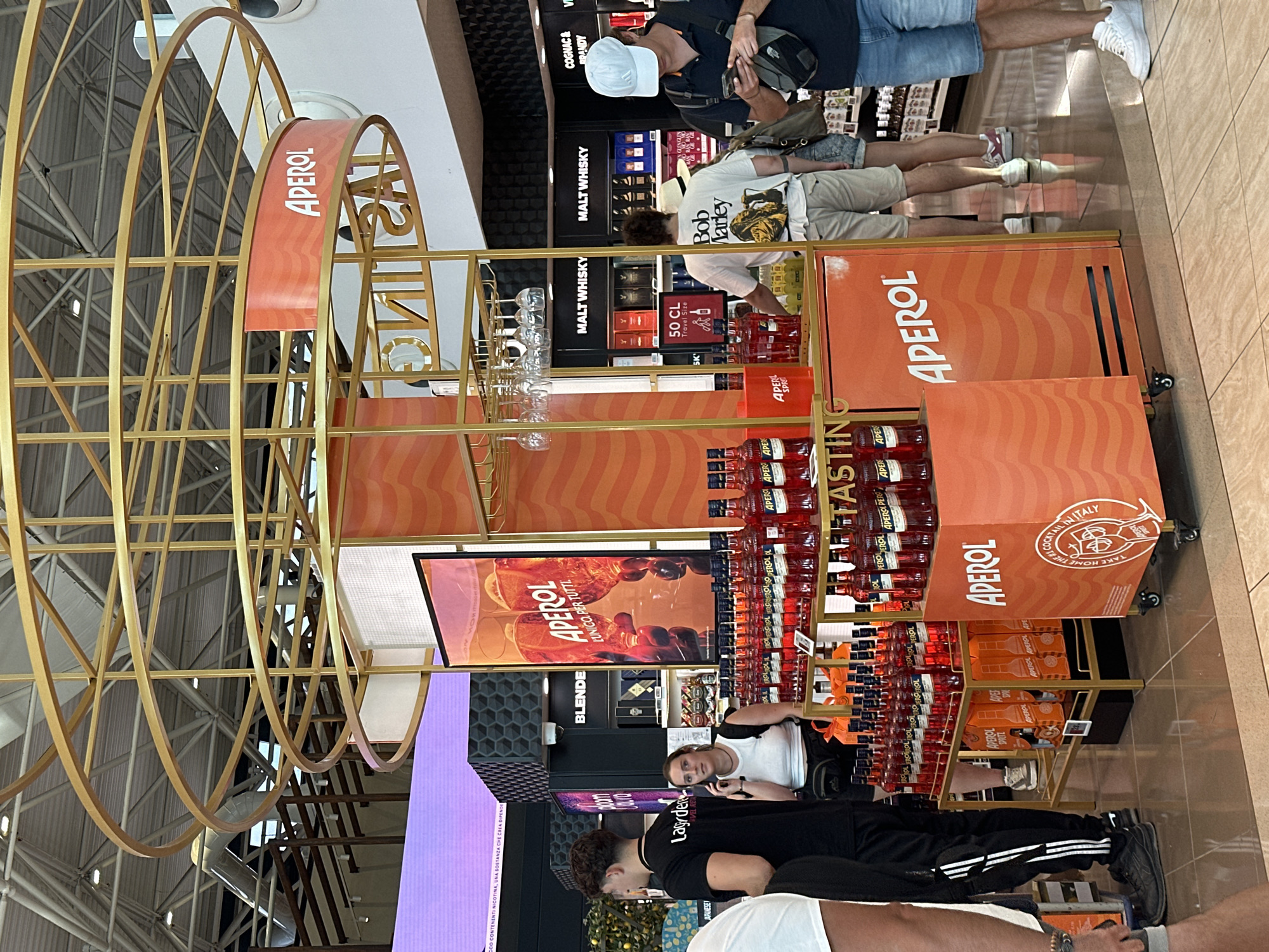 This image depicts an indoor commercial setting, likely an airport duty-free shop in Fiumicino, Italy, given the presence of brands like Aperol and various liquors. The foreground is dominated by a brightly lit orange display stand featuring numerous bottles of Aperol. The stand has a gold metal frame and is adorned with the "Aperol" logo and wavy orange patterns. A large sign on the stand reads "TASTING" in yellow lettering. Behind the display, shelves stocked with liquor bottles are visible, with prominent labels for "MALT WHISKY."
Several individuals are present. In the left mid-ground, a man wearing a black t-shirt with white stripes down the sleeves and black pants stands facing towards the right. Next to him, a woman in a white tank top and black shorts is talking on her mobile phone. To the right of the Aperol stand, a man wearing a white t-shirt with a "Bob Marley" graphic and beige shorts is looking at his phone. Another person, partially visible to the right, wears a blue t-shirt, denim shorts, and a white cap, also engaged with their phone.
The environment includes a high ceiling with exposed industrial elements and lighting fixtures. A large, vibrant purple screen is visible in the background on the left. The floor is tiled. The overall impression is a commercial space designed for product promotion and sales.