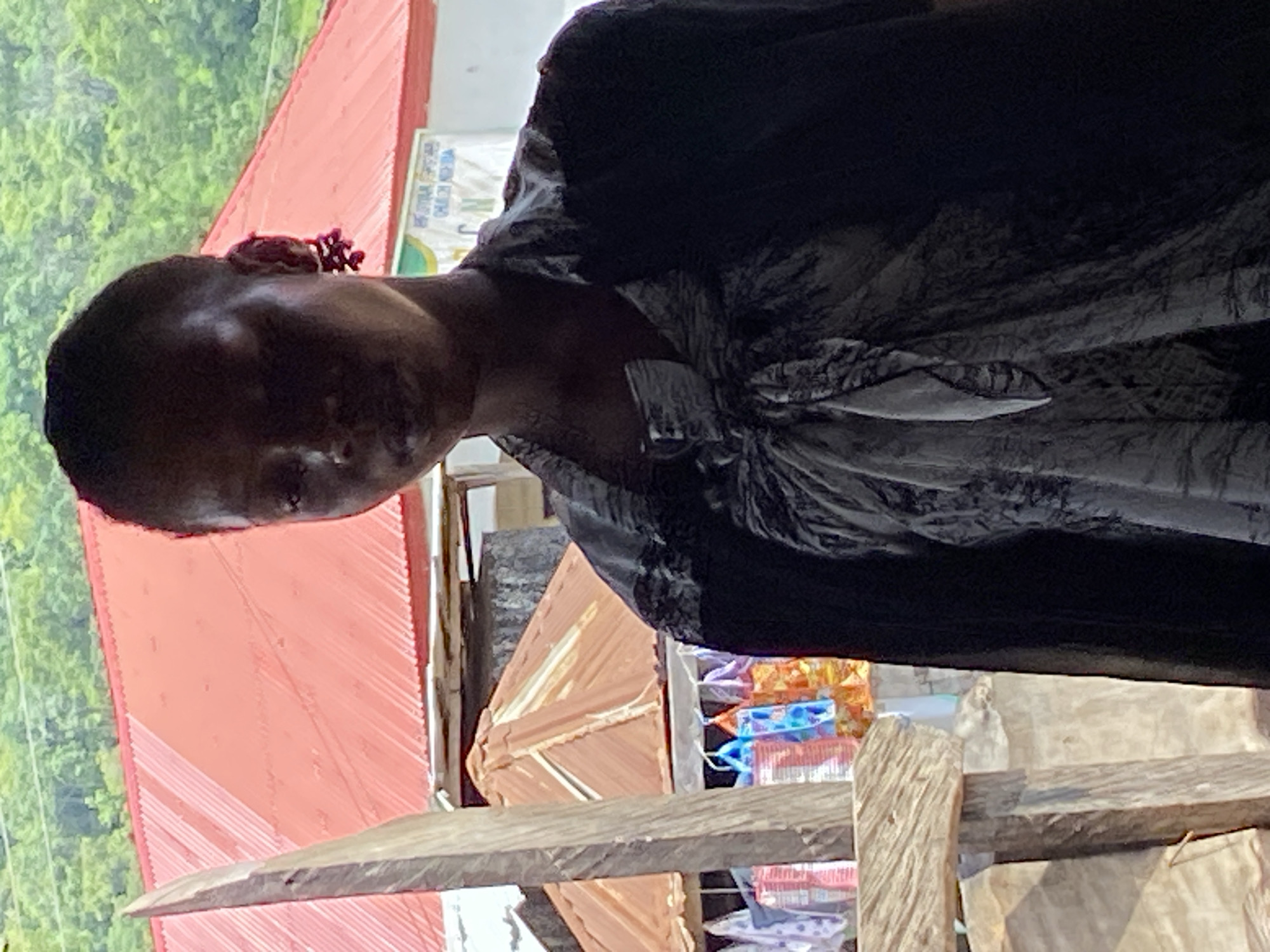 This is a portrait of a person in Mararaba, Nigeria. The subject is a person with dark skin and short hair, wearing a dark, patterned garment. The person appears to be looking down and to the left. In the background, there is a red roofed structure and green foliage, suggesting an outdoor or semi-outdoor setting. The lighting suggests it is daytime. There are some objects visible in the background that appear to be stacked items, possibly merchandise or building materials. A sign with text is partially visible on a white surface behind the person.