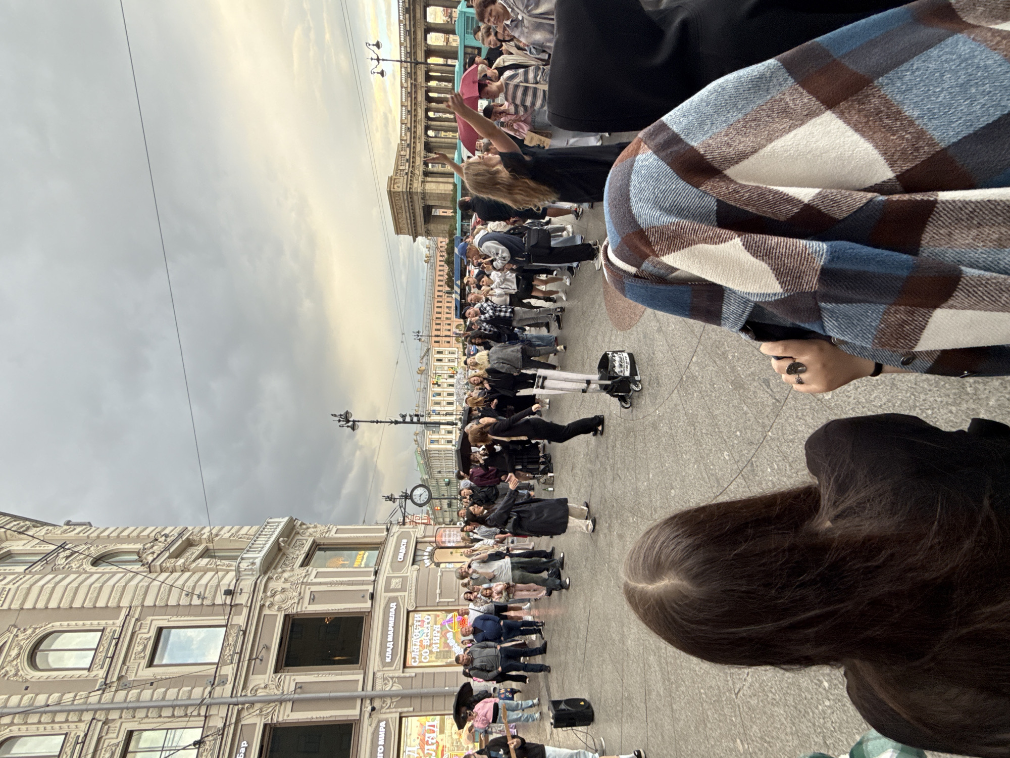The image depicts an outdoor scene in Saint Petersburg, Russia, likely during the day given the overcast sky and diffused light. A crowd of people is gathered on a paved area, possibly a plaza or street, in front of ornate buildings with European architectural styles. The sky is predominantly gray and cloudy, suggesting a cool or mild day.
The central focus appears to be a group of performers or entertainers. One individual, a woman with blonde hair, is in the middle of the frame, her arms raised and her body in a dynamic pose, suggesting she is part of a performance or dance. She is wearing a black dress. Other people are standing or sitting, watching the activity. Some individuals are holding umbrellas, indicating the possibility of light rain or as a stylistic choice. There are also what appear to be small, wheeled devices, possibly part of the performance or for personal mobility.
In the foreground, the back of a person wearing a plaid jacket is visible, obscuring a portion of the scene. Another person with long dark hair is also in the foreground.
Visible text includes signs on the buildings. One sign in Cyrillic appears to read "КЛАД МАРМЕЛАД" (Klad Marmelad), which translates to "Treasure Marmalade". Another sign reads "СЛАДКОГО СО-ВКУСО МИРА" (Sladkogo So-vkusa Mira), meaning "Sweet Taste of the World".
The overall atmosphere seems to be one of public gathering and entertainment, with an air of casual observation from the onlookers. The architecture of the buildings, with their detailed facades and numerous windows, strongly suggests a European city setting, and the specific Cyrillic text confirms the location in Russia.