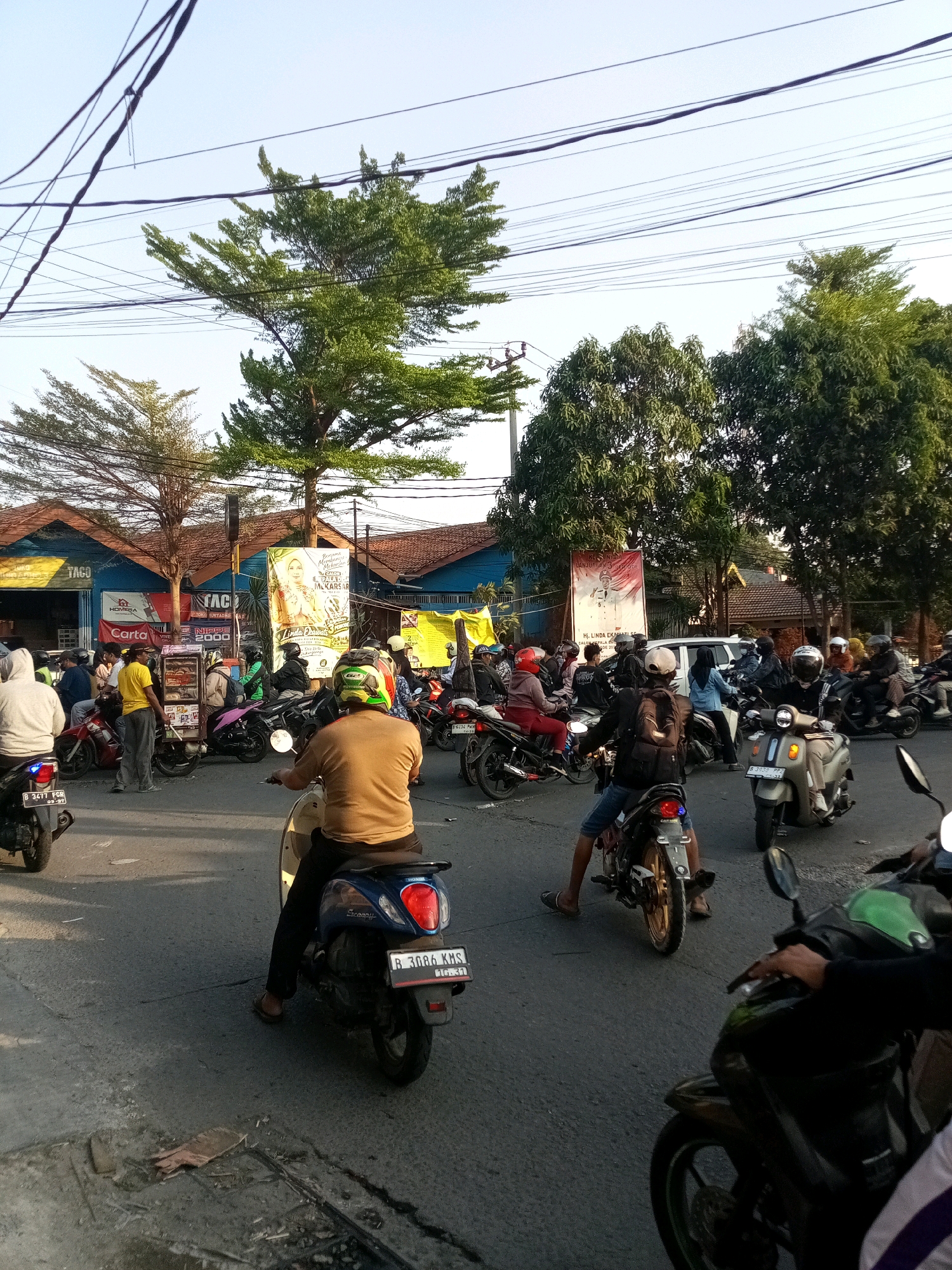 In Tambun, Indonesia, on a paved road, a large group of people are riding motorcycles amidst trees and buildings. The sky is clear and blue, with power lines crisscrossing overhead. Buildings with red-tiled roofs and blue and yellow exteriors line the street. Several large banners are displayed, one featuring a woman in a headscarf. The motorcycles are of various makes and colors, and many of the riders are wearing helmets. The scene appears to be a busy intersection or street where traffic is congested. Some people are standing by the roadside, while others are riding. A man in a yellow shirt is standing next to a small cart. The overall impression is one of a bustling urban environment in Indonesia.