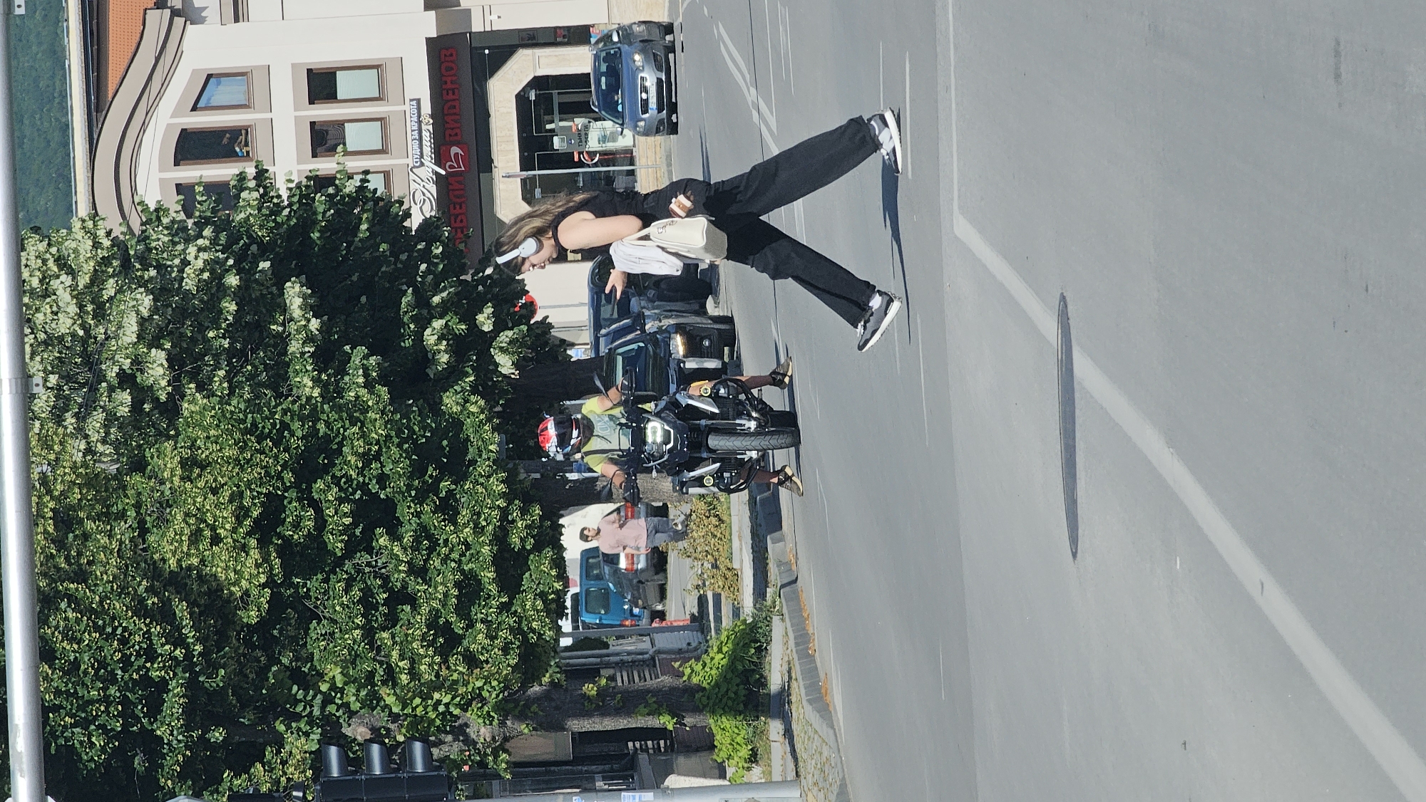 A female pedestrian wearing headphones and black pants crosses a street in Karlovo, Bulgaria. She is looking at her phone and carrying a white bag and a white jacket over her left shoulder. A male motorcyclist wearing a helmet and a yellow shirt is stopped at the intersection, facing the camera. He is on a black motorcycle with a large headlight. Behind the motorcycle, there are several cars stopped at the intersection. A man in a pink shirt is walking on the sidewalk in the background. Buildings line the street, including a shop with a sign that says "МЕБЕЛИ В ВИДЕНОВ". The scene is illuminated by sunlight, casting shadows on the road.