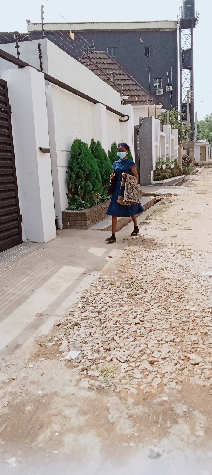 An image taken in Kaduna, Nigeria, depicts a female subject walking on an unpaved, uneven dirt path with scattered light-colored gravel and debris, moving from right to left across the frame. She wears a dark blue, short-sleeved dress, dark shoes, a blue head covering, and a light blue surgical mask. She carries a dark handbag on her left shoulder and a patterned fabric bag or item draped over her right forearm.
To the left of the subject, a tall, white perimeter wall with an electric fence system on top runs parallel to the path. A dark brown gate is partially visible on the far left. A security camera is mounted on this wall. Below the wall, three conical green evergreen shrubs are planted in a brick planter box. Further along, another section of a white wall with decorative architectural elements and an electric fence is visible.
In the background, a multi-story, dark grey or black building with multiple windows and mounted air conditioning units stands prominently. A lower building with a brown-tiled roof is situated in front of it. A tall, dark metal structure supporting a black water tank is visible on the far right. The sky is bright but appears hazy or overcast. Shadows from an unseen structure are cast on the ground to the left of the subject.