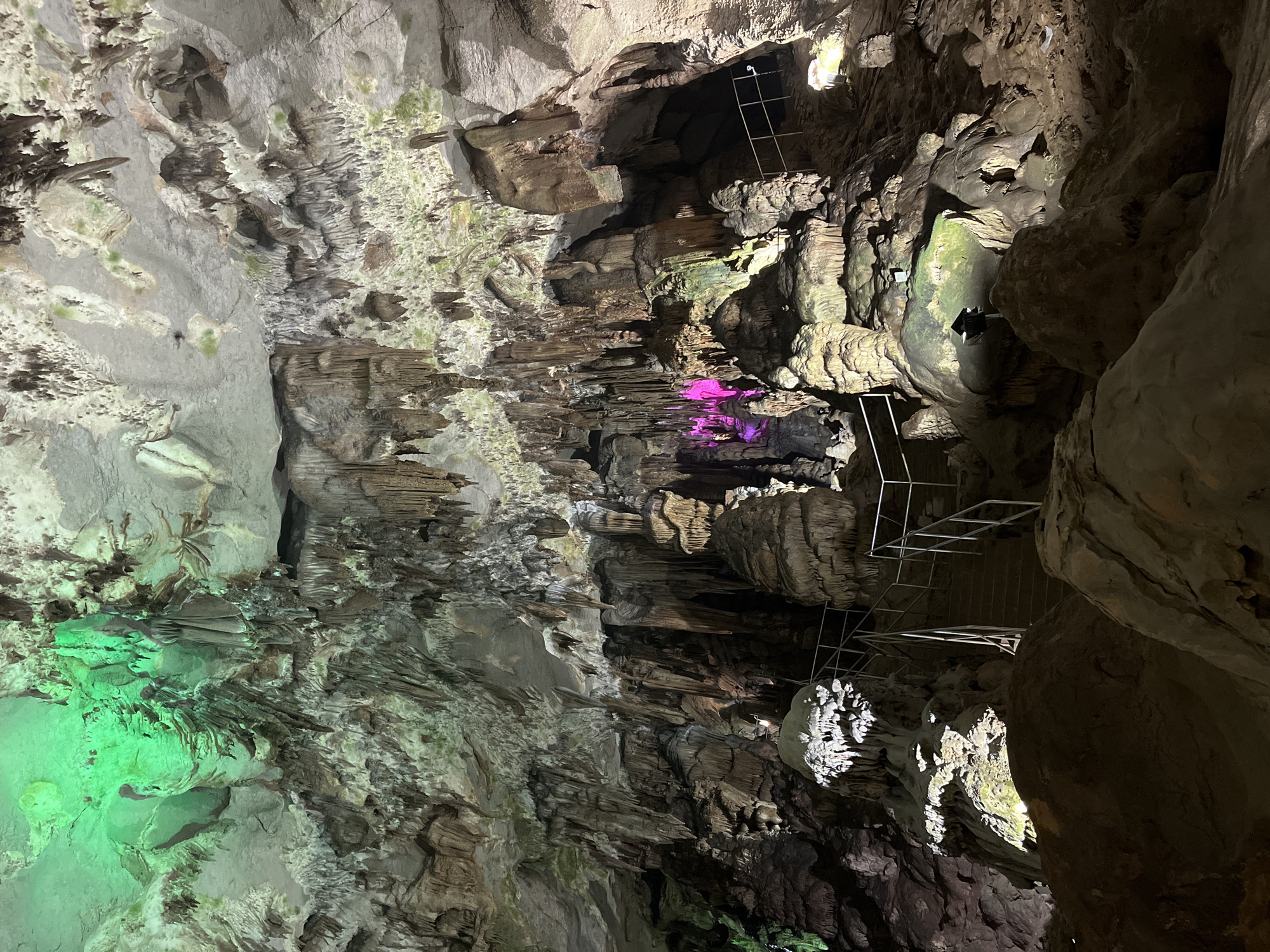An interior view of a large cave is shown, illuminated by artificial colored lights, including green and pink. Stalactites hang from the ceiling, and stalagmites rise from the floor, forming intricate formations. A metal railing and steps are visible, indicating a path for visitors. The cave walls are rough and textured, with some areas showing moss or mineral deposits. The scene captures the dramatic natural beauty of the cave environment.