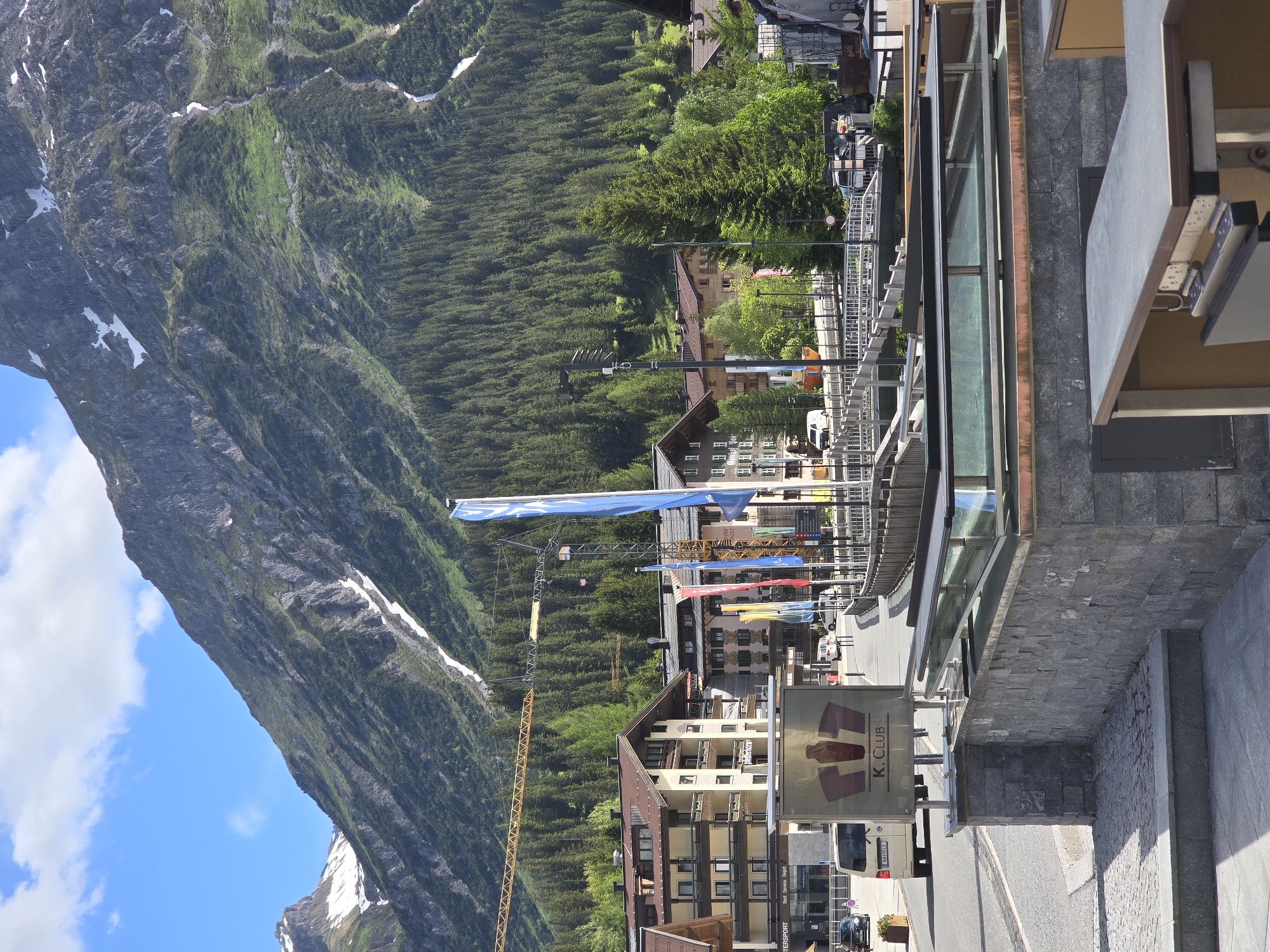 The image shows a view of the town of Lech, Austria, with a large mountain in the background. The mountain is covered in trees at the base and has patches of snow near the summit. In the foreground, there are several buildings with traditional Austrian architecture, including balconies and wooden elements. A road runs through the town, with a few cars and a white van visible. Several flags, including blue ones, are planted along the road. There is also a construction crane in the middle ground. A sign for "K. CLUB" is prominently displayed on a metal pole. A stone wall with glass panels is in the immediate foreground. The sky is blue with some white clouds.