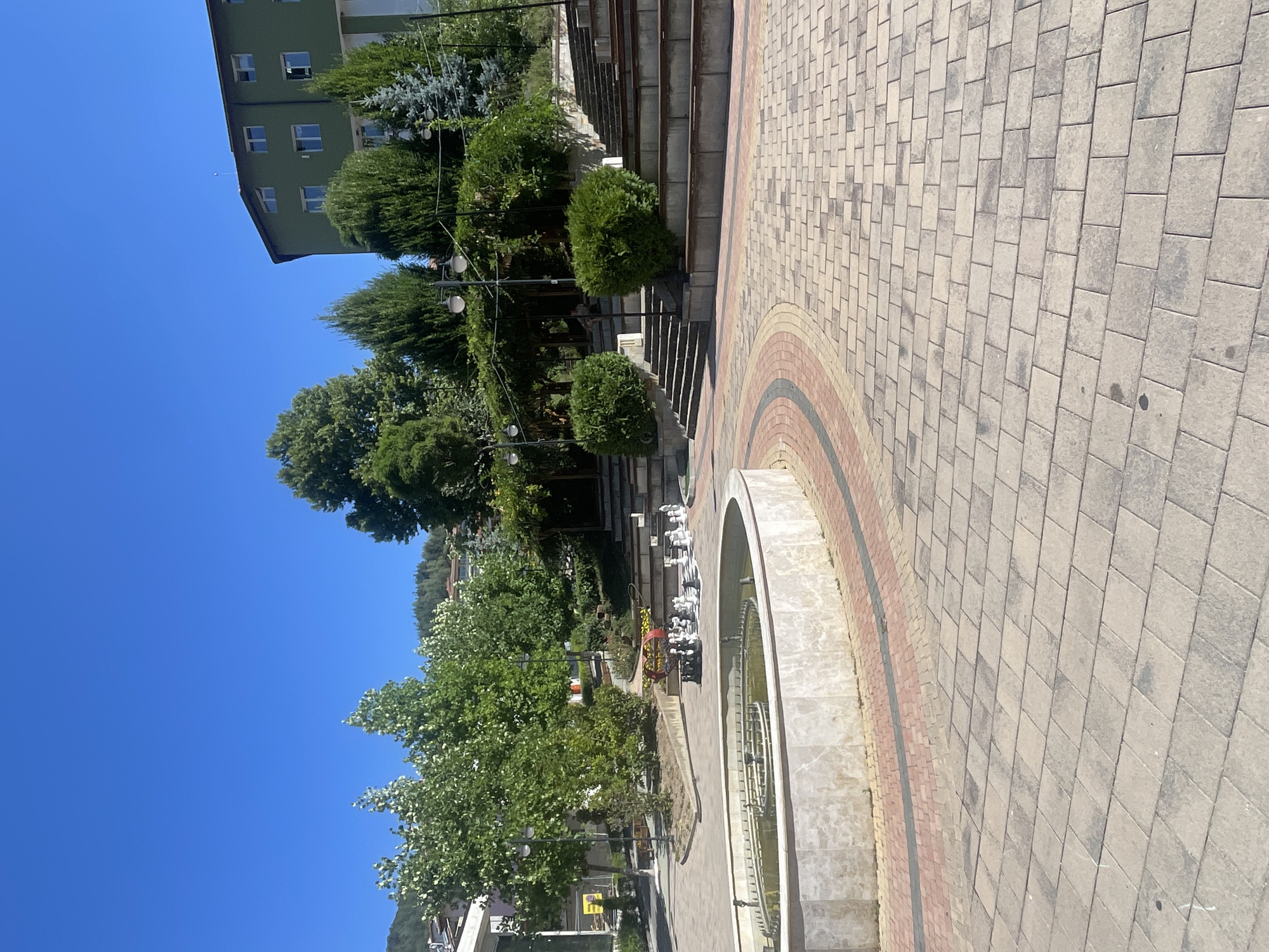 The image depicts an outdoor public space in the city of Banite, Bulgaria, featuring a paved plaza with a circular fountain in the foreground. The fountain has a stone basin with a marble-like finish and a tiered inner structure. The plaza is paved with interlocking rectangular bricks in shades of grey, beige, and red.
Behind the fountain, a large, circular outdoor chess set with white and black pieces is set up on the paved ground. To the right of the chess set, there are tiered seating areas constructed from concrete and wood, resembling an amphitheater. Several lush green trees and shrubs are interspersed throughout the scene, adding to the natural ambiance.
In the background, a multi-story green building with numerous windows is visible. The sky is clear and bright blue. The scene appears to be a well-maintained public area, likely a park or a town square, designed for relaxation and recreation.