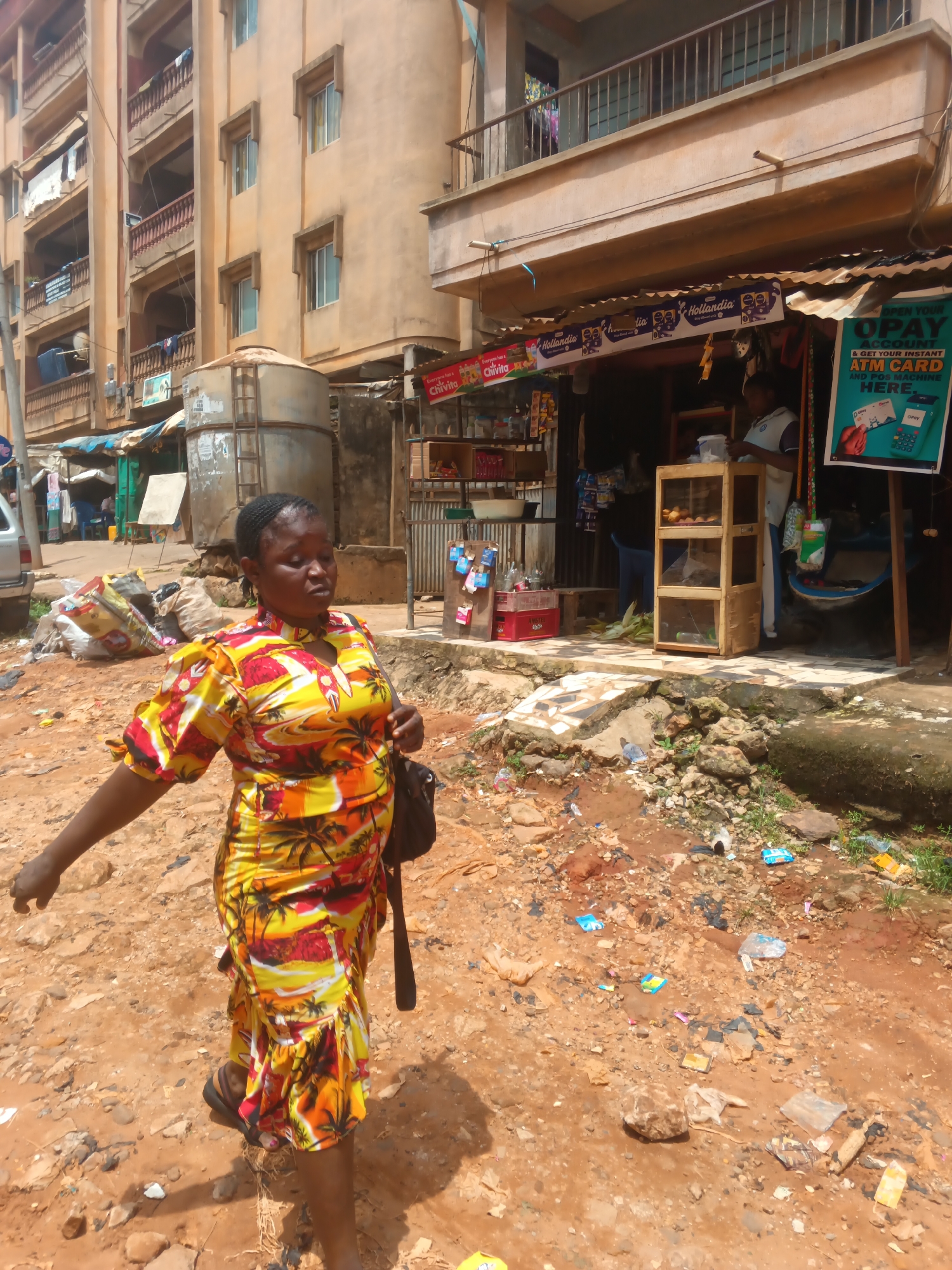 A woman in a vibrantly patterned dress walks across a littered, dusty street in Onitsha, Nigeria. The street is unpaved and uneven, with scattered debris including plastic bags, wrappers, and stones. In the background, a multi-story building with balconies and laundry hanging from them dominates the scene. Adjacent to the building is a small shop with an awning and shelves stocked with goods. A large metal water tank is visible to the left of the shop, and a parked car is partially visible on the far left. A poster advertising OPay services is displayed prominently on the front of the shop. The woman, carrying a dark bag over her shoulder, is moving from left to right across the frame, her gaze directed slightly downward and to the side.