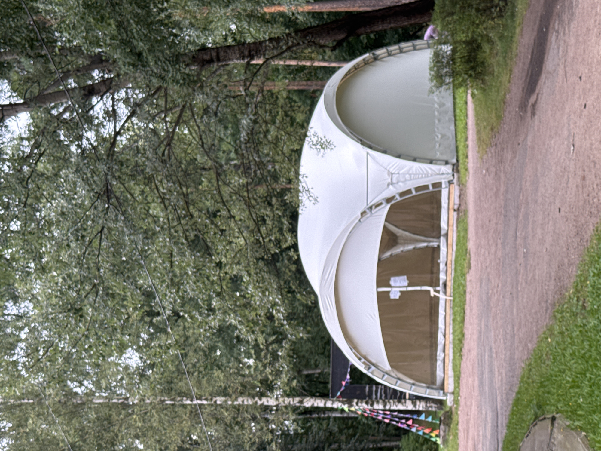 A white tent structure with arched openings is situated in a wooded area. The tent has a light brown interior visible through the openings. Colorful triangular flags are strung on a pole to the left of the tent. A dirt path leads towards the tent, with grass and trees surrounding the scene. The sky is overcast, and the lighting suggests it is daytime. The location is an outdoor public space.