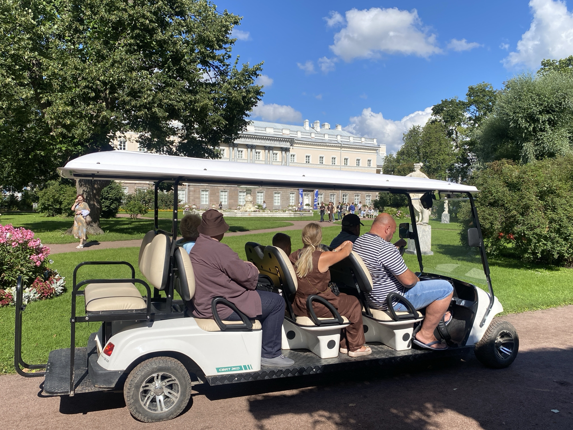 In Korpikiulya, Russia, a white electric golf cart, identified by the visible text "СВЯТ ЭКО" and "SE 62", travels along a paved path in a park on a sunny day. The cart carries five individuals: a man driving in a striped t-shirt and denim shorts, a woman with blonde hair in a brown outfit, a person in a brown hooded garment and dark pants, another person in a brown hooded garment, and a person in the far back of the cart. The park features lush green grass, mature trees with full canopies, and a large, multi-story building with classical architectural elements in the background, suggesting a historical estate or palace. A woman in a patterned dress walks on the grass to the left of the cart, and a display of pink flowers is visible near the cart's front left. The sky is blue with scattered white clouds.