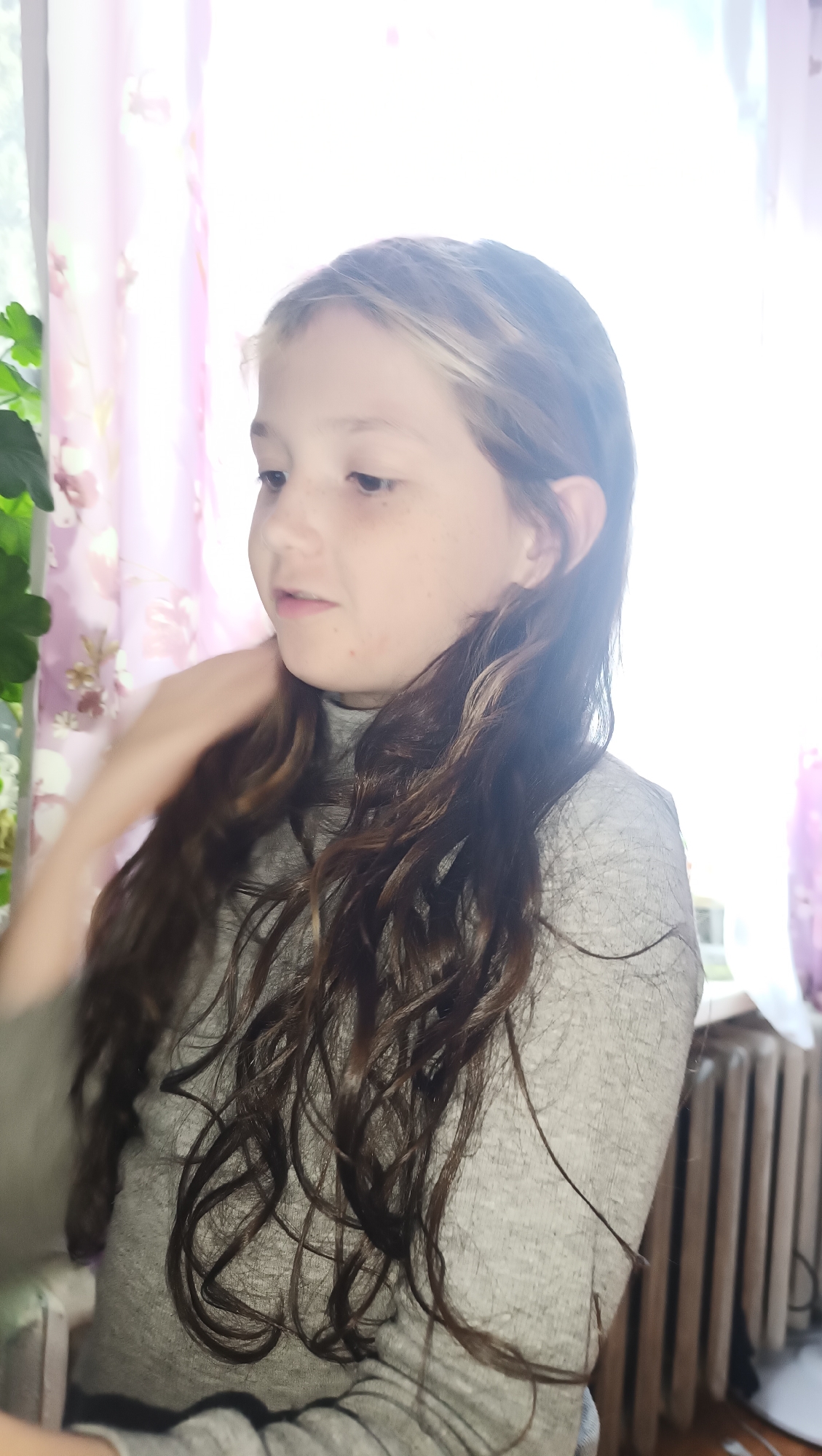 The image shows a young girl with long, wavy brown hair, looking to her left. She is wearing a grey turtleneck shirt. Her right hand is near her neck, with her fingers touching her hair. The background is blurred, with hints of pink floral curtains and a bright light source, suggesting a window. To the right, a radiator and a wooden floor are partially visible. There is no indication of the city of Karsha, Russia, in the image.
