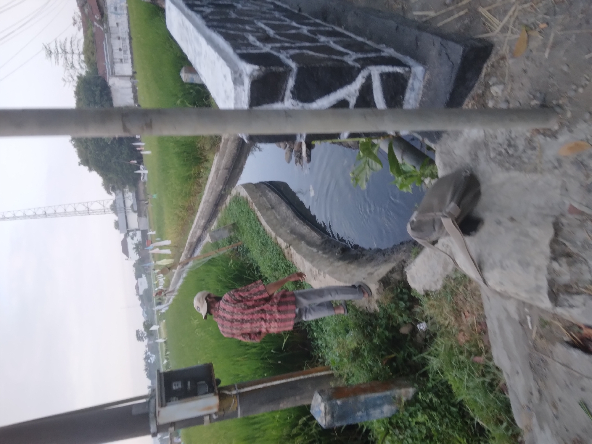 The image depicts an outdoor scene in Desa Pamijen, Kecamatan Sokaraja, Indonesia, with a focus on a person walking near a rice field and an irrigation canal. The person, wearing a red and black checkered shirt, grey pants, and a light-colored baseball cap, is walking along the edge of a concrete structure next to a dark water-filled canal. To the left of the person is a tall utility pole with a metal box attached to its base. The rice field stretches into the background, dotted with small white flags on poles. In the middle ground, a concrete bridge with a dark stone facade painted with white markings spans the canal. Green vegetation is visible along the banks of the canal and in the foreground. The sky is overcast. A brown bag lies on the concrete in the foreground, to the right of the person.