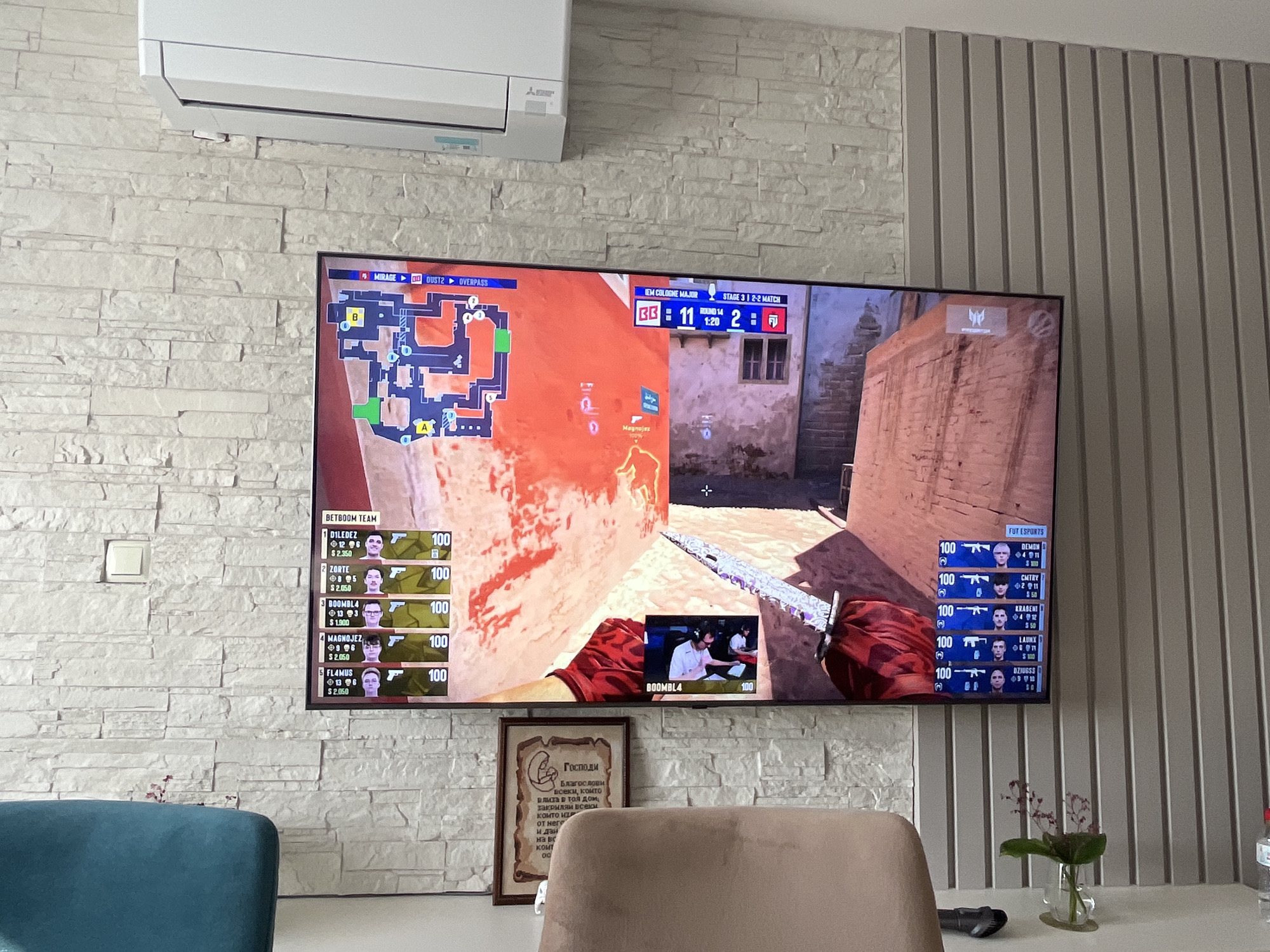 The image depicts an indoor scene with a large television displaying a video game, likely Counter-Strike, set in the city of Varna, Bulgaria. On the wall behind the television, there is a white air conditioning unit. The wall itself is covered with white decorative stone tiles, and to the right, a section of the wall features vertical grey panels.
The television screen shows a competitive gaming match, with a minimap in the upper left corner and player statistics displayed on both the left and right sides of the screen. The game appears to be in progress, with team scores and player names visible. A small inset screen in the lower right of the main game display shows a live view of some of the players. Below the television, a framed religious text is hung on the wall.
In the foreground, a portion of a teal-colored chair is visible on the left, and a beige chair is in the center and right. A small vase with a plant and some flowers sits on a table in front of the beige chair. A bottle of water and a black device, possibly a remote control, are also on the table.
The overall impression is a modern living space adapted for entertainment, with a focus on watching and possibly playing video games.
