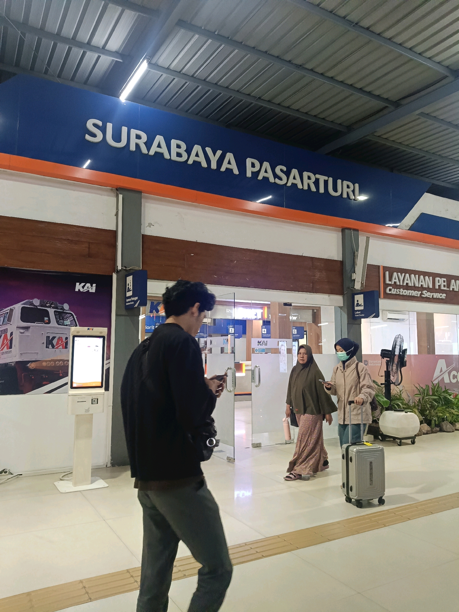The image shows a view inside a train station in Surabaya, Indonesia. A large blue sign with white lettering reads "SURABAYA PASARTURI". In the foreground, a person wearing a black jacket and grey pants walks away from the camera, looking at their phone. To the right, two women are walking towards a customer service area. One woman is wearing a green hijab and a long patterned skirt, carrying a pink water bottle. The other woman, wearing a beige jacket and blue jeans, is wearing a face mask and pushing a grey suitcase on wheels. Behind them, there is a customer service counter with the words "LAYANAN PELANGGAN Customer Service" and a standing fan. To the left, a self-service kiosk with a screen displaying information is visible, along with a large advertisement featuring a train engine with the KAI logo. The environment appears to be an indoor, covered area of the station with a corrugated metal roof and tiled flooring.