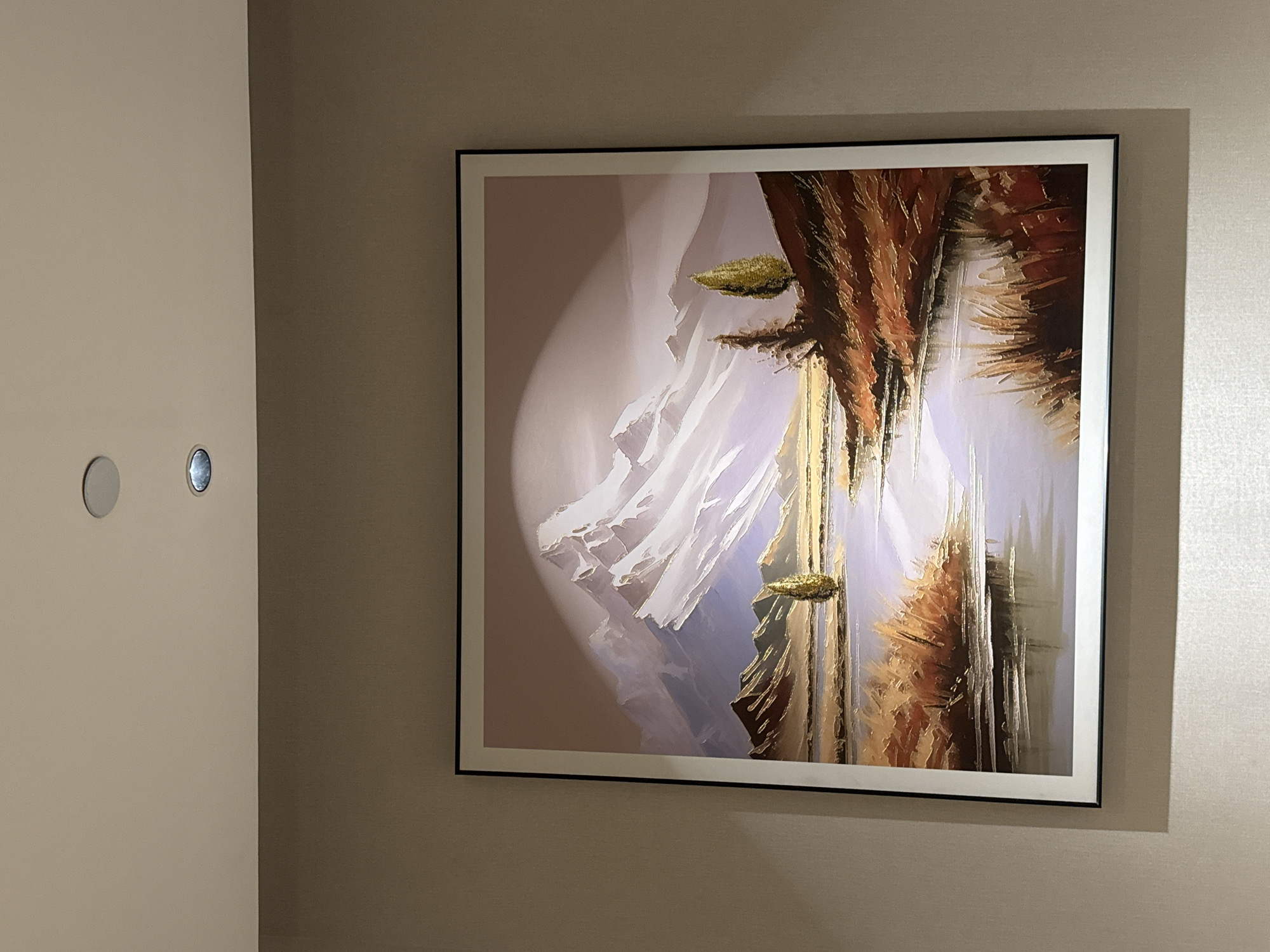 A framed painting of a mountain landscape is mounted on a light beige wall. The painting depicts snow-capped mountains in the background, a body of water in the middle ground reflecting the sky, and a shoreline with dry grass and trees. The overall color palette is muted, with shades of white, blue, brown, and purple. Two recessed ceiling lights are visible above the painting. The image was taken in Chaoyang District, China.