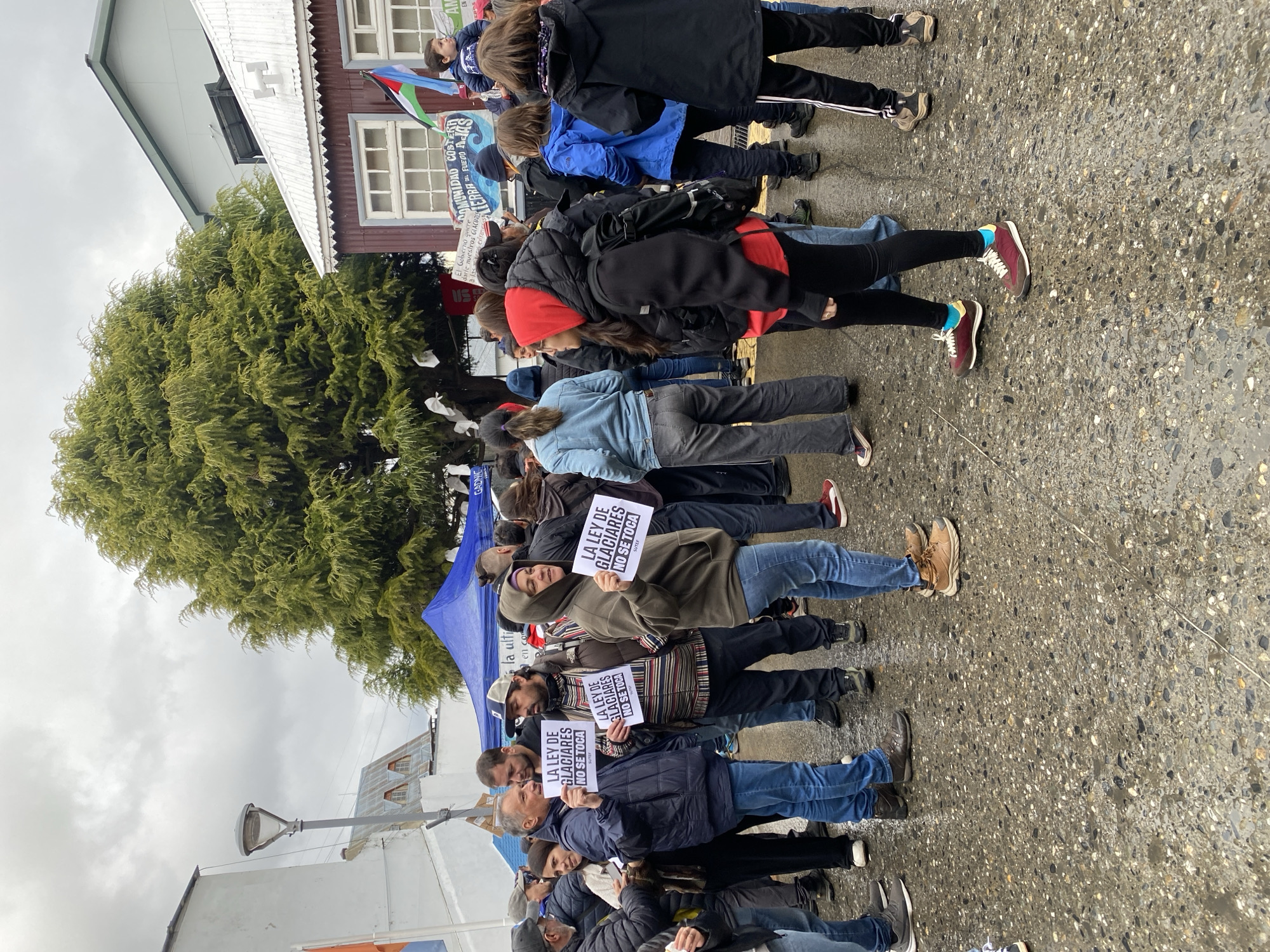 The image captures a protest in Ushuaia, Argentina. A group of people stand close together, holding signs that read "LA LEY DE GLACIARES NO SE TOCA" (The Glacier Law is Not Touched). They are standing on a wet, speckled pavement near a tree. Buildings are visible in the background. The sky is overcast, suggesting a cloudy day.