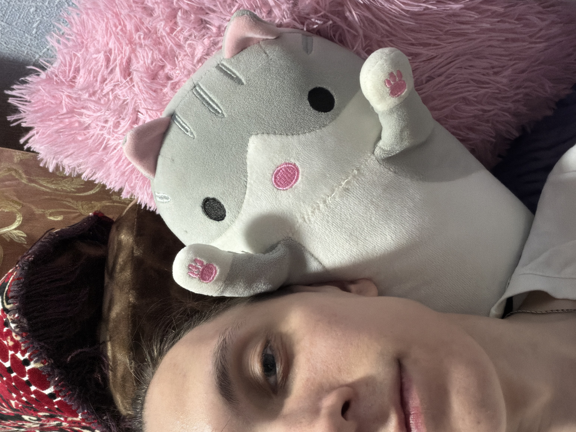 In a close-up, selfie-style shot, a person's face and upper body are visible, positioned horizontally within the frame. They are lying down, with their head tilted towards the left side of the image. A gray and white plush cat toy rests beside them, its paws extended upwards. The cat is positioned on top of a fluffy pink pillow, which occupies the right side of the frame. In the background, a dark-colored patterned fabric is visible. The overall environment appears to be an indoor setting, possibly a bed or sofa. The lighting suggests an indoor environment, with soft illumination. The image does not provide any information about the location within a city.