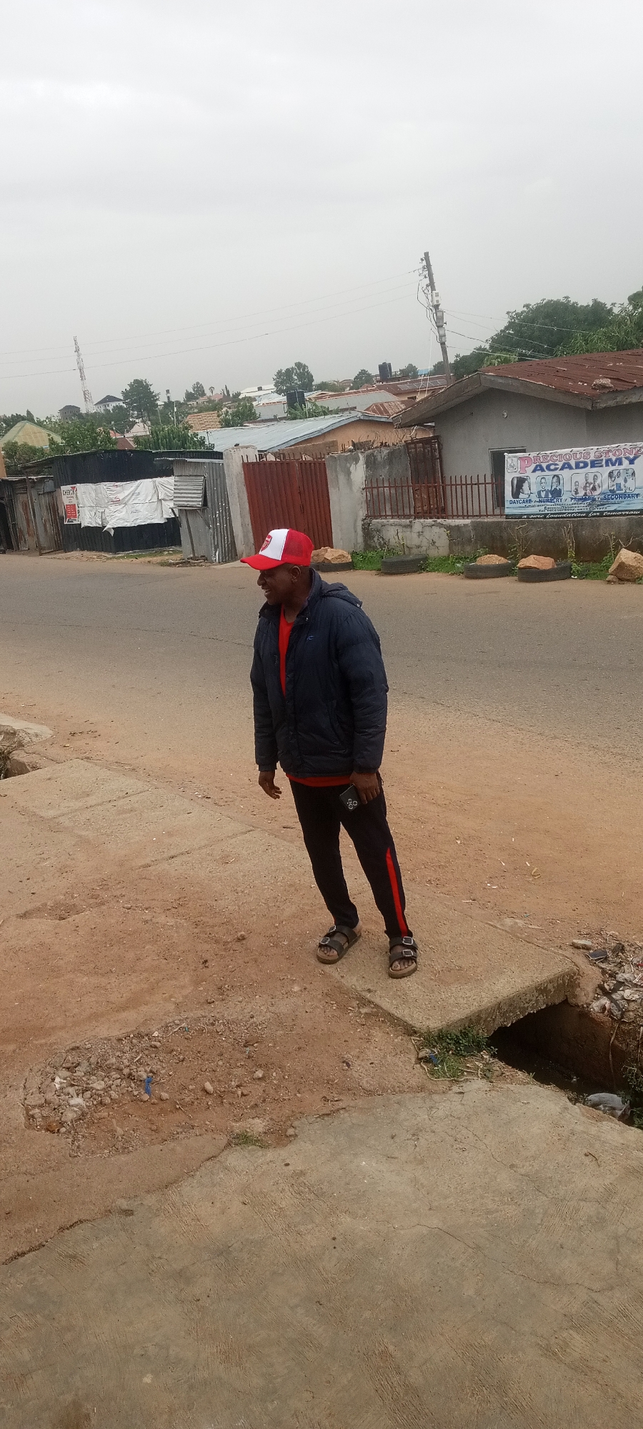 A middle-aged man wearing a red and white cap, a navy blue jacket over a red shirt, black pants with a red stripe, and sandals, stands on a concrete structure at the edge of a road in a residential area. The man is facing slightly to the left, looking towards a building across the road. The road appears to be paved, but with some dirt and gravel on the edges. Behind the man, there are houses with corrugated iron roofs and fences. One building has a sign that reads "PRECIOUS STONE ACADEMY". Overhead power lines are visible against an overcast sky.
