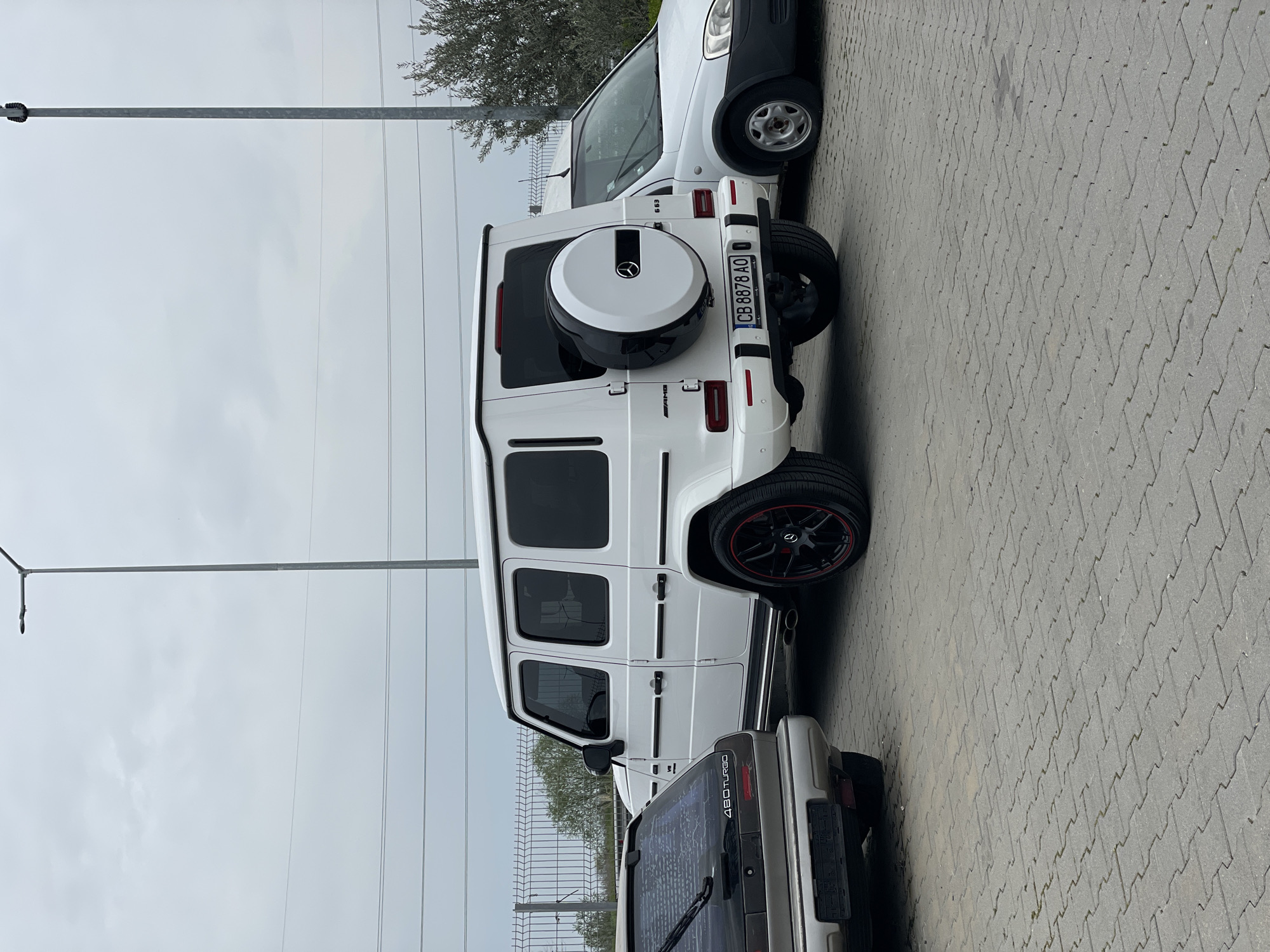 The image shows a parked white Mercedes-Benz G-Class SUV with a Bulgarian license plate CB 8878 AO. It is parked on a paved surface alongside other vehicles, including a white van and a grey car with a "480 TURBO" sticker. The setting appears to be an outdoor parking area. A light post and a metal fence are visible in the background. The sky is overcast. The location is indicated by the license plate, which is Bulgarian and might be near Asenovgrad.