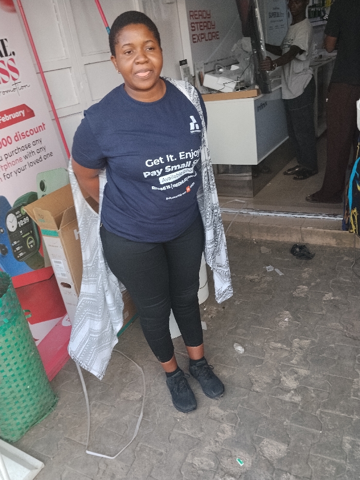 An outdoor scene in Fagge C, Nigeria, depicts an adult female standing in the foreground, facing the viewer with a slight smile. She has a medium-dark complexion and short, dark hair. She wears a dark blue short-sleeved t-shirt with white text that reads, "Get It. Enjoy. Pay Small Small." and "Available in February From N15k REGAL." below a white logo. Black leggings and black athletic shoes with black socks complete her attire. A white and gray patterned shawl or lightweight garment is draped over her shoulders.
Behind her, to the left, are various objects: a tall green woven plastic basket, stacked cardboard boxes (one appearing to be a "Hisense" product box, another red), and a promotional sign with text partially visible, mentioning "PROMOTION," "February," "000 discount," and showing images of smartwatches and possibly smartphones.
To the woman's right, a glass storefront entry reveals the interior of an electronics shop. Inside, an adult male wearing a light-colored t-shirt and dark pants is visible, appearing to adjust or carry a large, flat, dark object (possibly a television or display screen). Display counters are visible, one labeled "Infinix." A red and white sign on the back wall reads, "READY STEADY EXPLORE." The lower legs and feet of a second individual are partially visible to the far right, near the shop entrance.
The ground is a textured, light gray pavement with an interlocking pattern, showing minor debris including a white strap and small pieces of litter.