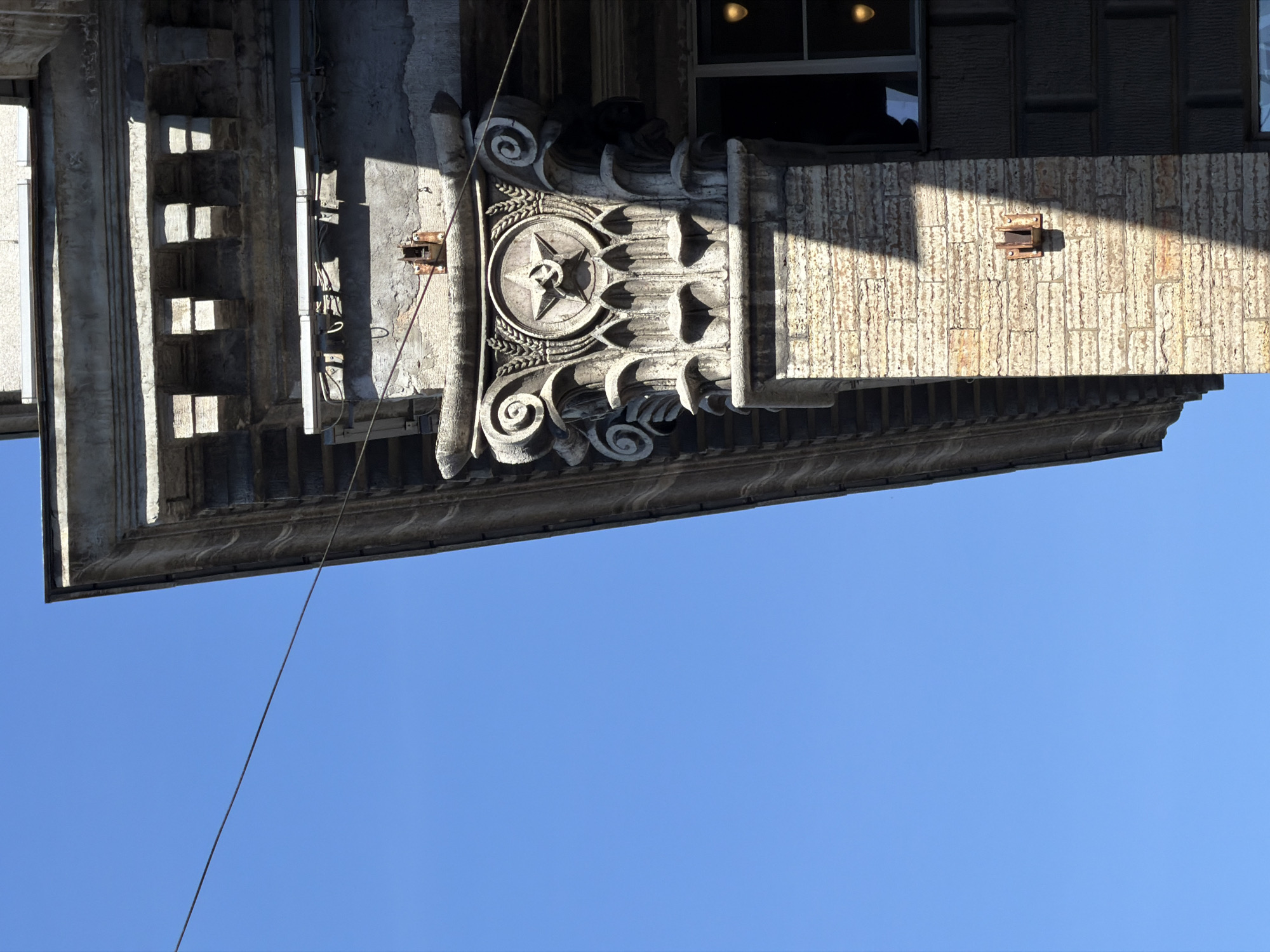 The image presents an upward-looking view of the ornate stone facade of a building against a clear blue sky. The architectural style blends classical elements with specific motifs, potentially indicating a historical period like the Soviet era in Saint Petersburg, Russia.
A central feature is a highly detailed, carved stone column or pilaster. It prominently displays a circular medallion with a deeply etched five-pointed star at its core, flanked by stylized wheat or laurel leaves. Above and below this central motif, intricate scrollwork designs, characteristic of volutes, adorn the stone. The stone shows signs of weathering and age.
To the right of this decorative column, the building's exterior transitions to a section constructed from light-colored, rectangular blocks, possibly travertine or rough-hewn stone, which is brightly illuminated by direct sunlight. Sharp shadows highlight the texture of these blocks. A small, rusted metal bracket is attached to this block wall. Further up and to the right, a dark-framed window reflects internal lights.
On the left side of the ornate column, a plainer section of the facade is visible, featuring vertical fluting and recessed rectangular panels, also marked by distinct shadows. Some areas show weathered concrete or stucco and exposed utility conduits or wires.
The lower portion of the image is dominated by a vast expanse of cloudless blue sky. A thin, dark wire or cable runs diagonally across this sky from the upper left towards the bottom right. The scene is brightly lit, indicating a sunny daytime. There are no people or discernible text visible in the image.