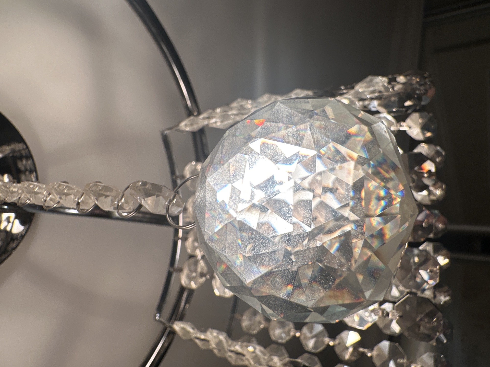 This is a close-up, eye-level shot of a clear, faceted glass orb, likely a decorative element of a chandelier or light fixture. The orb is round and appears to be made of many small, flat surfaces that catch and refract light, creating rainbow-like glints across its surface. It is suspended from a thin, metallic loop or ring.
Strands of smaller, clear glass beads, also faceted, are attached to the orb and trail away from it, connected by thin metal rings. These strands are arranged in a cascading fashion, suggesting they are part of a larger decorative piece. The background is out of focus and consists of muted, neutral tones, with a soft light source illuminating the main subject. The lighting suggests an indoor setting, possibly a room with ambient light or a light fixture that is on. There is no discernible text, people, or weather conditions visible. The overall impression is one of sparkle and delicate craftsmanship.