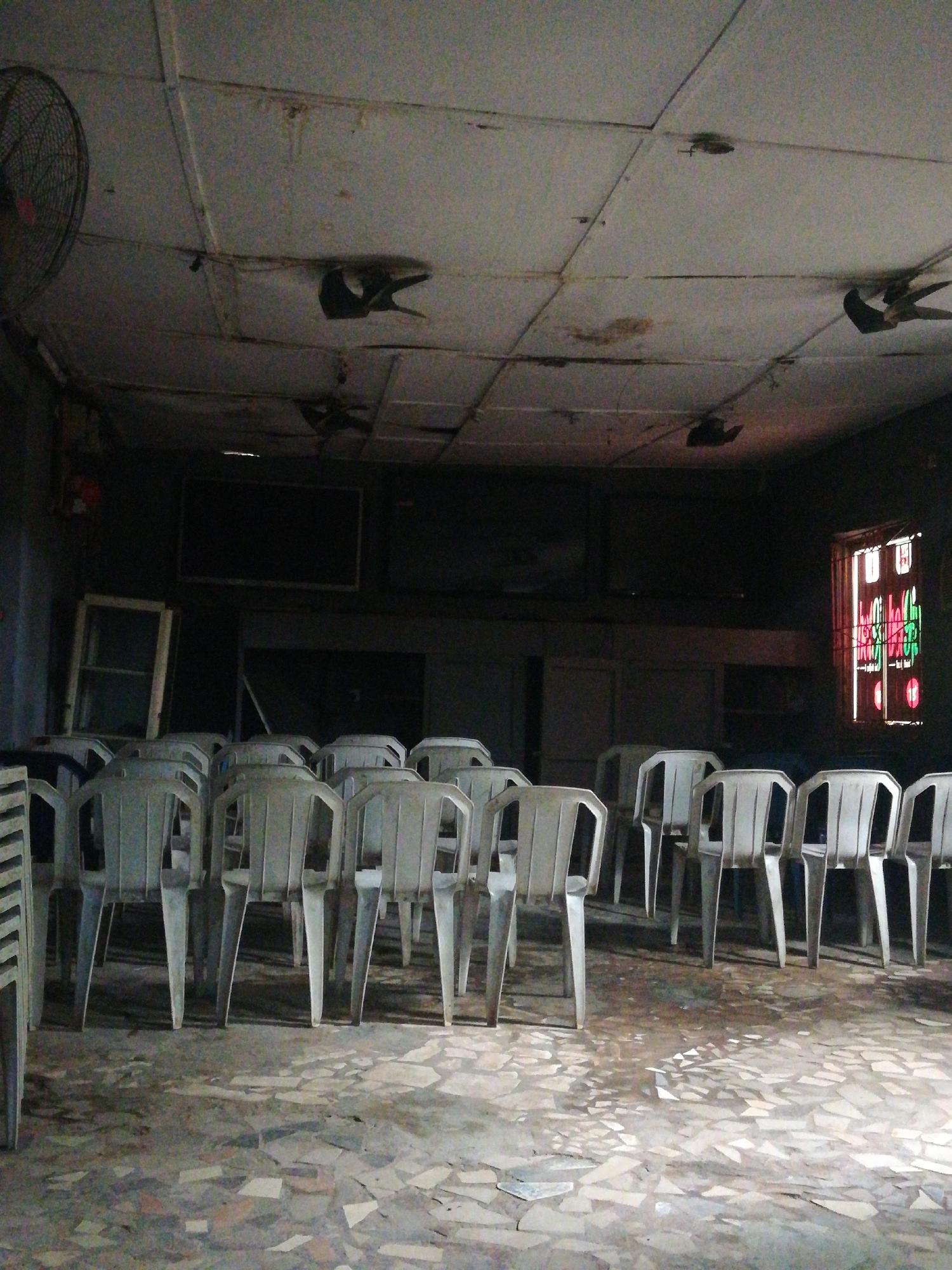 The image shows a dimly lit interior, possibly in Lagos, Nigeria, based on the style of the window and ceiling fixtures. Rows of white plastic chairs face a dark area with obscured objects, possibly TVs or screens. A stained glass window is on the right side of the image. The ceiling is low and has visible staining and damage, with a fan on the left. The floor has a fragmented tile pattern. There are no visible people.