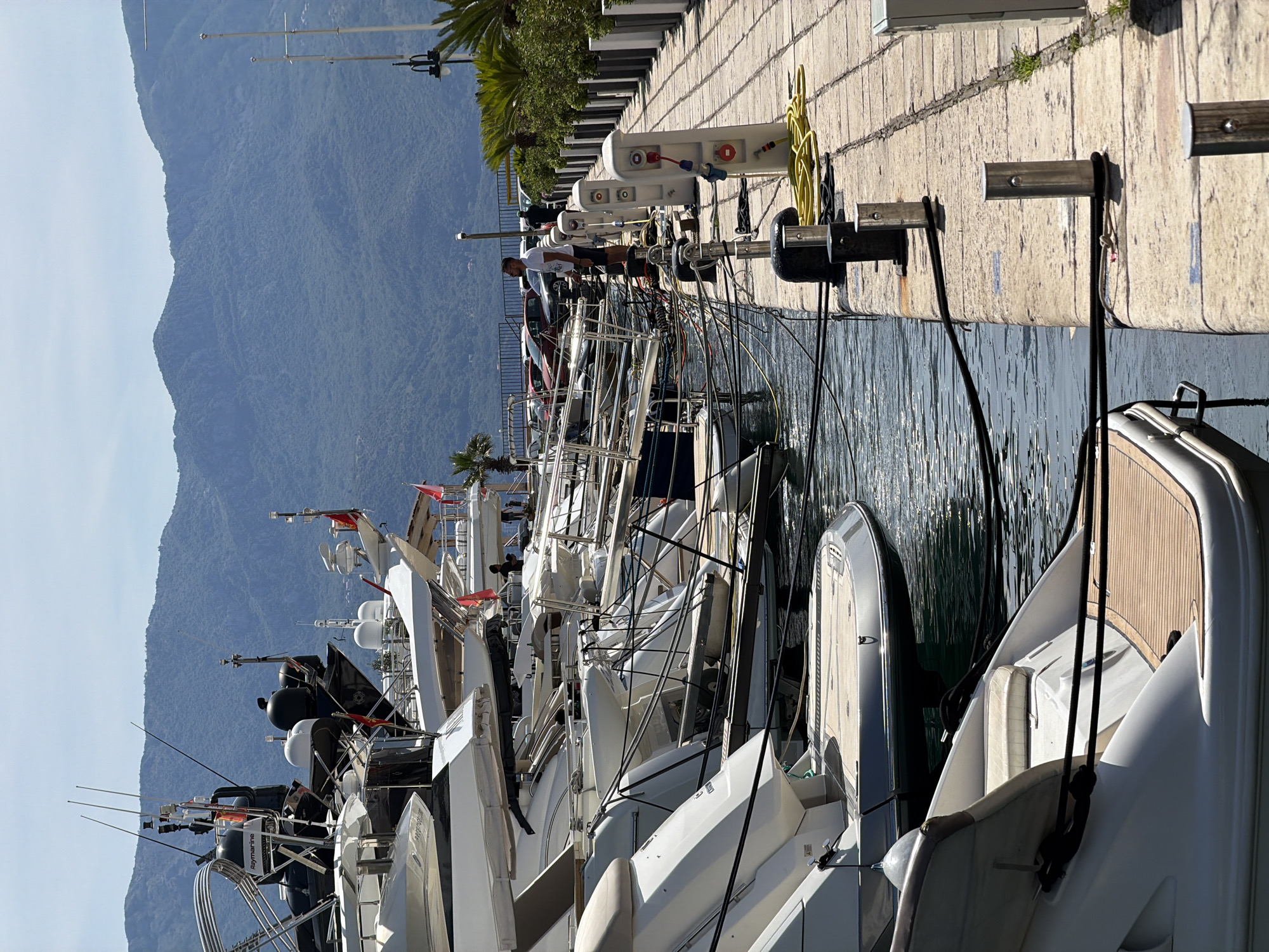 The image depicts a marina in Budva, Montenegro. Multiple yachts are docked along a concrete pier. The pier is equipped with utility boxes and mooring fixtures. A man stands on the pier attending to a vessel. Mountains rise in the background under a clear sky. The water reflects the moored yachts.
