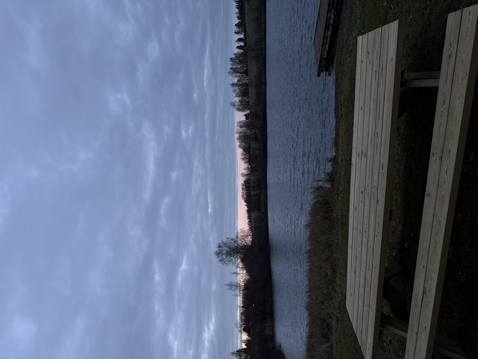 The image displays an outdoor scene at twilight, likely in a recreational area within or near Sand, Norway. The sky is overcast with gray-blue stratocumulus clouds, exhibiting a subtle pink-orange hue along the horizon line. In the foreground, a light-colored wooden picnic table, consisting of a rectangular top and two attached benches, is situated on short-cut green grass. A dark blue body of water, presumed to be a lake, occupies the midground, showing minor surface ripples. The shoreline immediately adjacent to the water features patches of dried tall grass and reeds on the left. On the right side of the water, a wooden dock, constructed from horizontal planks, extends a short distance into the lake. The distant shoreline is densely forested with a mix of bare deciduous trees and some evergreens, backed by gently rolling hills. No individuals, animals, or explicit actions are present within the frame. The scene is static, illuminated by low, diffuse light.