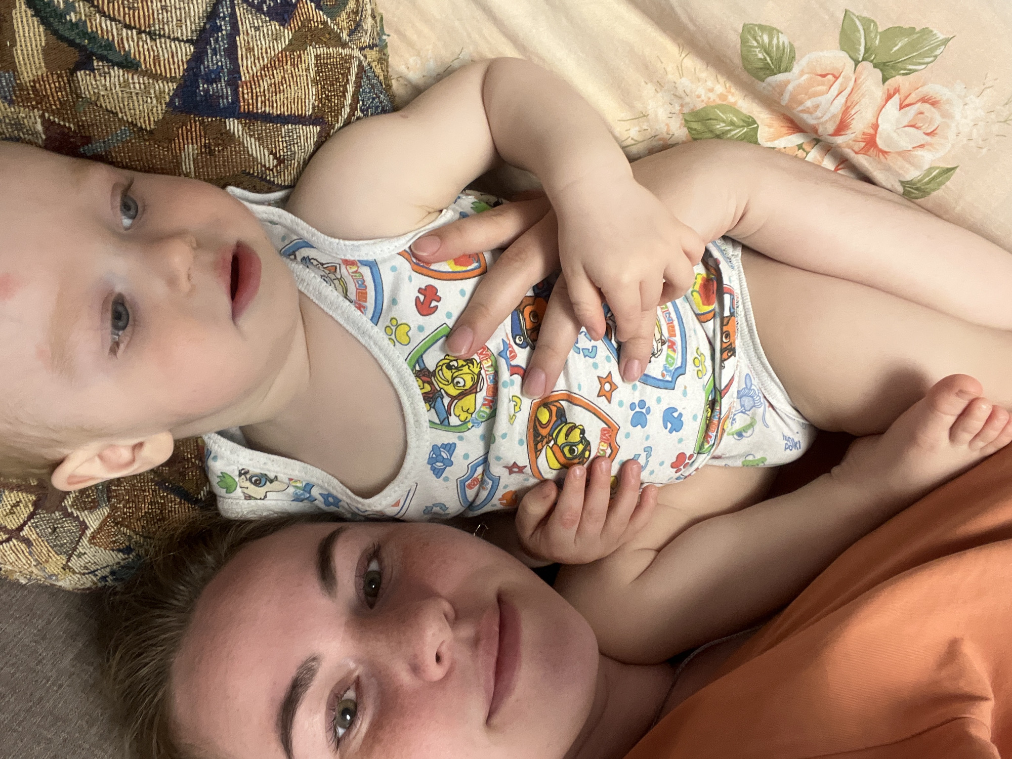 The image shows a woman and a young child lying on a bed. The woman is at the bottom of the frame, looking up towards the camera. The child is lying on top of her, facing the upper left corner. The child is wearing a white onesie with Paw Patrol characters printed on it. A hand is visible on the child's stomach. The bed has a floral pattern on the right side and a woven texture on the left. The woman is wearing an orange top. The location is the city of Chaikovsky, Russia.