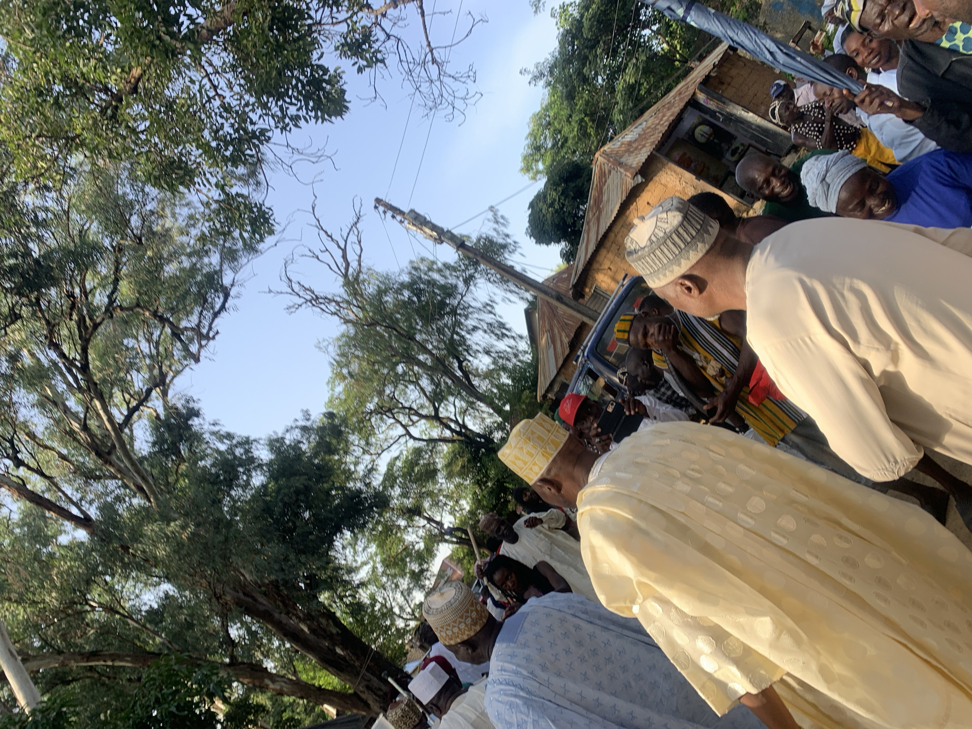 The image depicts a gathering of people, predominantly men, in what appears to be an outdoor, semi-rural setting in Jos, Nigeria. The subjects are dressed in traditional attire, including several individuals wearing kufi caps and flowing robes, suggesting a cultural or religious event. One person in the foreground is wearing a striking yellow robe with a subtle circular pattern. The group is clustered together, with some individuals looking towards the camera and others engaged in conversation or observing something.
The scene is characterized by abundant greenery from large trees with thick foliage overhead, dappling the ground with shade. A building with a corrugated metal roof and earthen walls is visible in the background, hinting at a residential or commercial area. Utility poles and wires also suggest the presence of infrastructure.
The time of day appears to be late afternoon or early evening, with the sunlight casting warm tones and long shadows. The sky is a clear, pale blue. The overall mood seems convivial, with smiles visible on several faces, suggesting a positive and perhaps celebratory occasion. The presence of an umbrella, held by one of the individuals, might indicate preparation for shade or a light drizzle. No visible text is present in the image.
