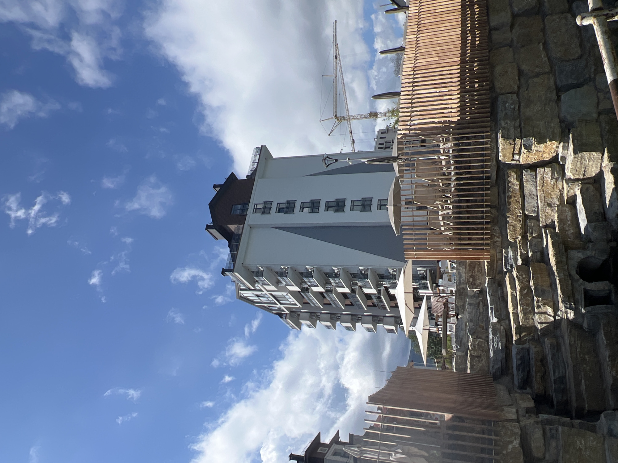 The image shows a modern multi-story building with balconies against a bright blue sky with scattered clouds. In the foreground, there is a terraced area constructed from large, rough-hewn stones, forming steps. A wooden fence with vertical slats runs along the top of the stone structure, enclosing an outdoor seating area with umbrellas and chairs. In the background, construction cranes are visible, indicating ongoing development. The scene appears to be a resort or residential complex. The location is identified as Yaremche, Ukraine.