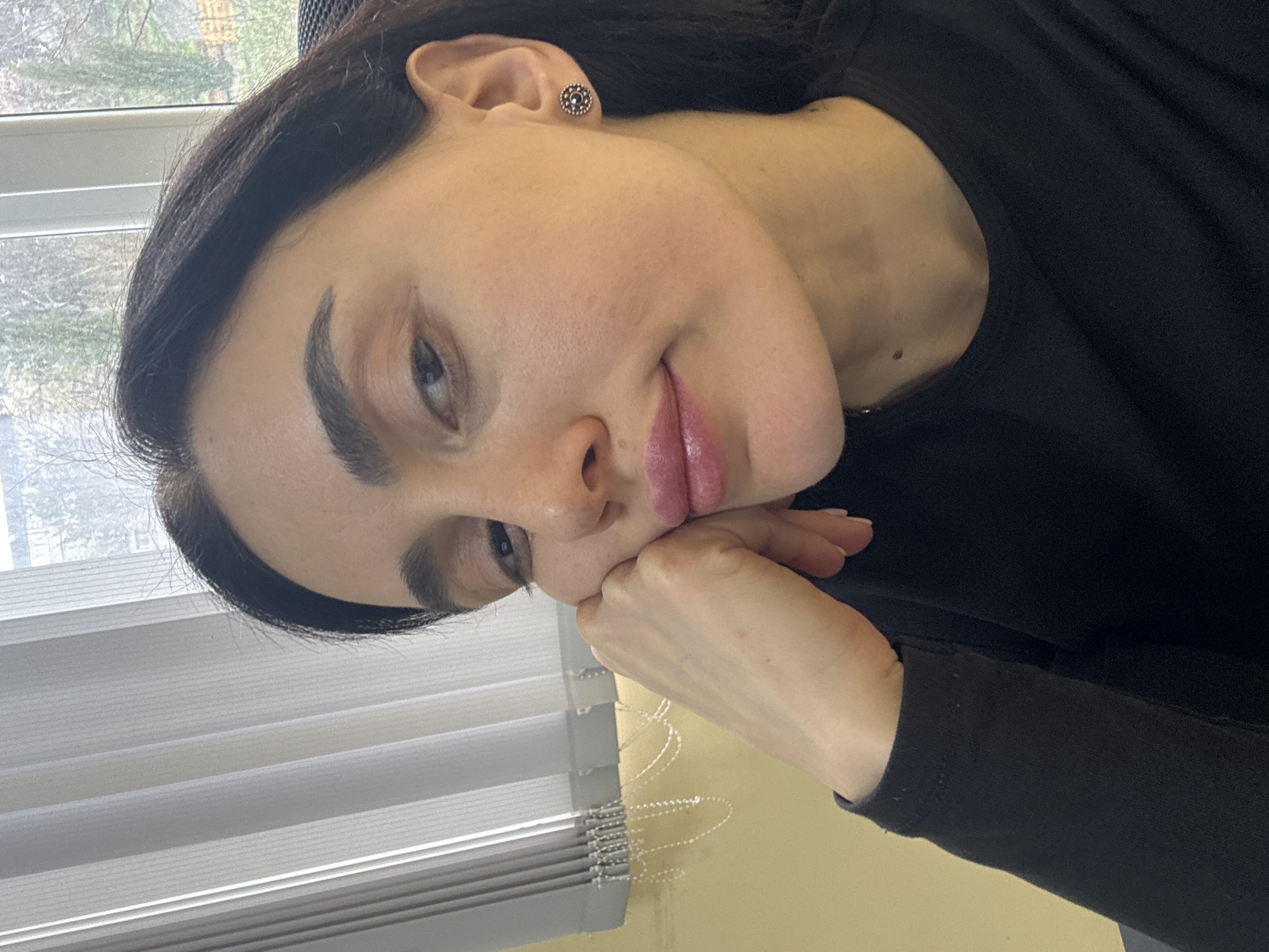 A woman with dark hair and light skin rests her face on her hand, looking directly at the camera. She is wearing a black long-sleeved shirt and a small earring. The background features a window with closed blinds, suggesting an indoor setting. Based on the architectural elements and general context, this scene could be occurring in a residential or office building in Tbilisi, Georgia, though no specific landmarks are visible to confirm the exact location.