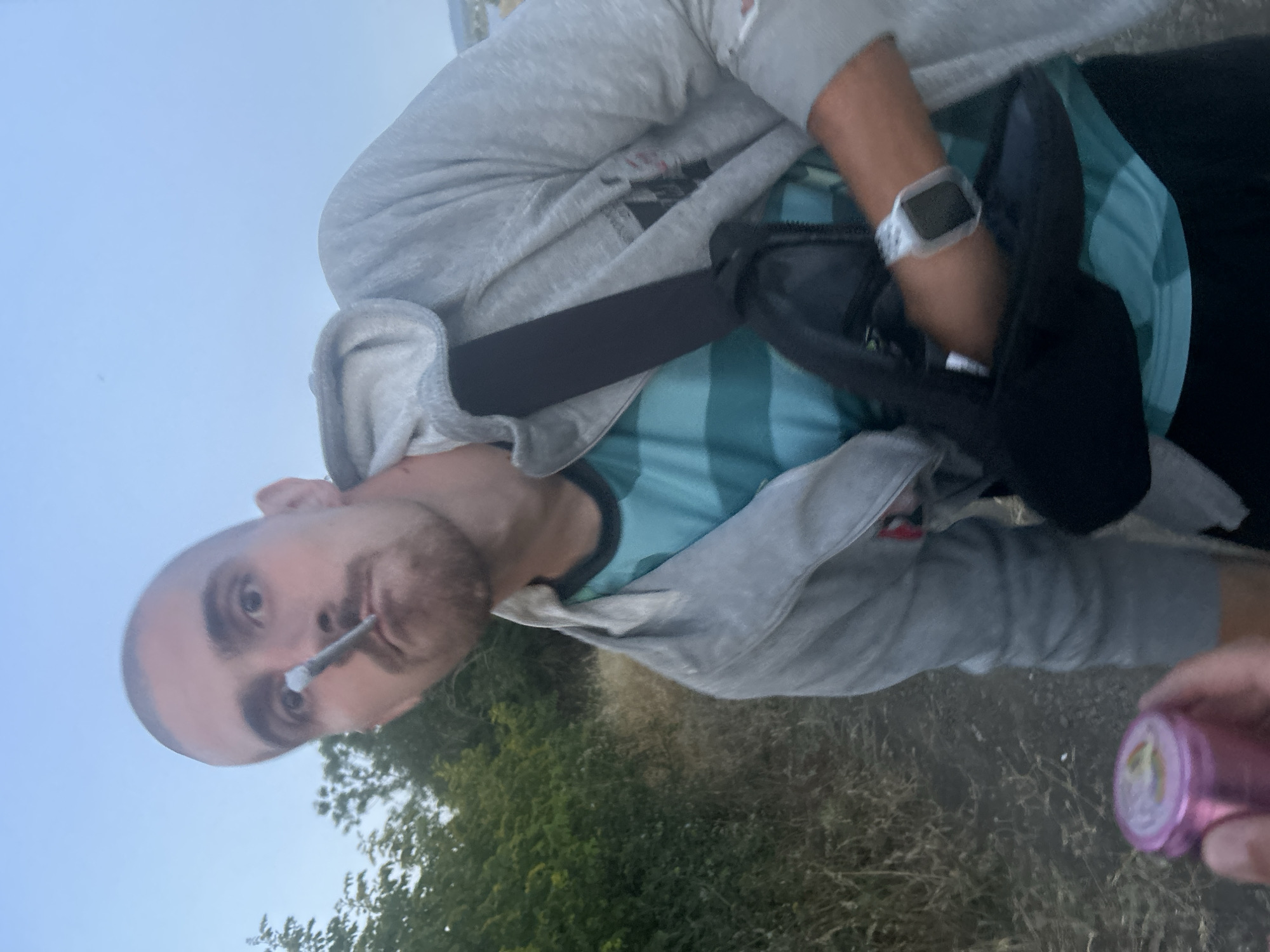 A person with a shaved head and a goatee is holding a smoking object in their mouth. They are wearing a grey hooded jacket over a teal and green striped t-shirt and black pants. A black bag is slung across their chest. The person is standing in an outdoor environment with dry grass and trees in the background. A pink cylindrical object, possibly a grinder, is being held in the foreground by another person, partially visible. The scene appears to be in the city of Burgas, Bulgaria, indicated by the presence of what looks like a distant coastline or bay in the background. The sky is overcast, suggesting it may be dusk or early morning.