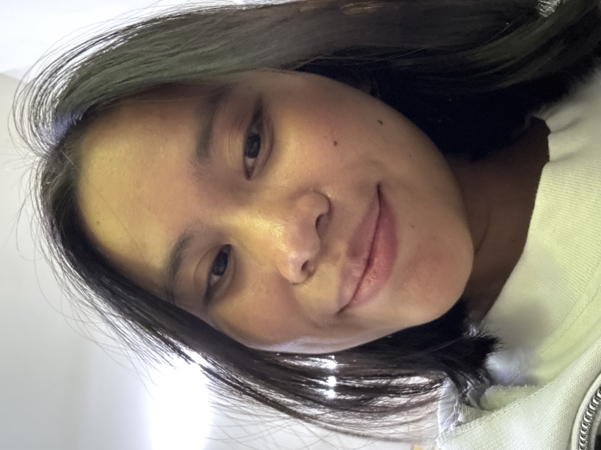 The image is a close-up, slightly angled selfie of a young East Asian woman. She has dark hair that is slightly tousled and frames her face. Her eyes are dark and she is looking slightly off-camera. Her lips are slightly parted, and she appears to be smiling softly. She has a small mole on her right cheek. She is wearing a light-colored hooded sweatshirt, part of which is visible at the bottom of the frame. The background is blurred and appears to be an indoor setting with bright lighting, possibly a wall or ceiling. The image quality is good, with clear focus on the woman's face. There is no discernible location in East Jakarta, Indonesia, or any specific environmental details that can be identified from the image.