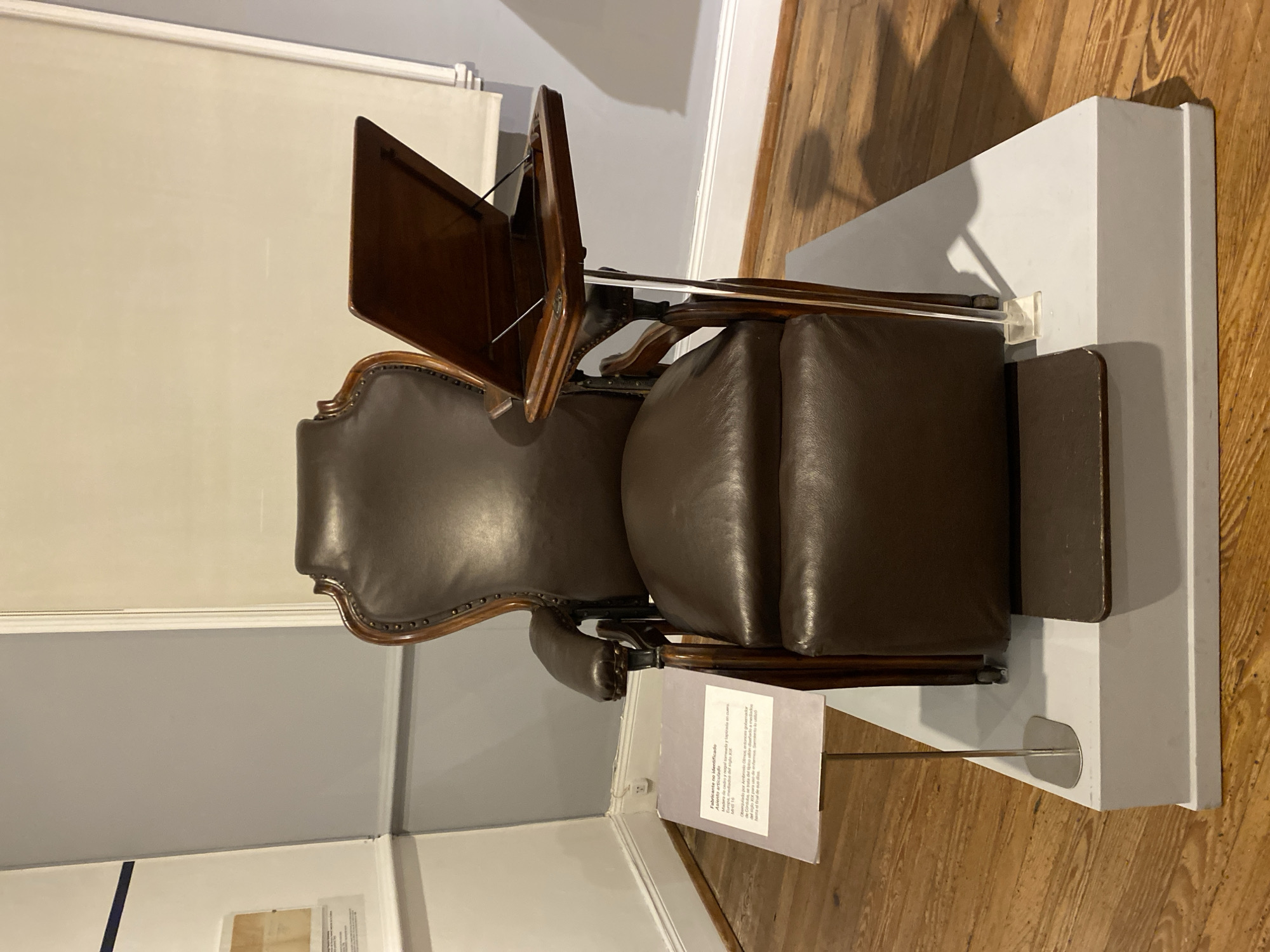 A top-down medium shot shows a brown leather chair displayed on a white platform, possibly in a museum or exhibit. The chair has a hinged writing surface attached to one arm. A sign, likely providing information about the chair, stands in front of it. The scene suggests a historical or antique furniture display, potentially located in the city of Buenos Aires, Argentina, based on the prompt. The environment appears indoors, with wooden flooring and neutral-colored walls.