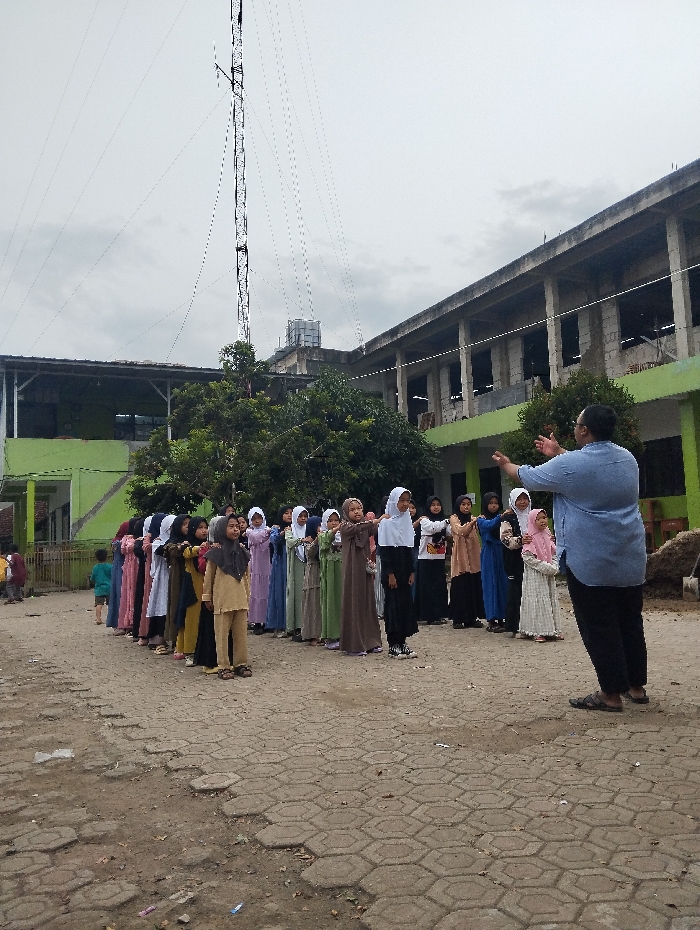 A group of approximately 25 young girls, all wearing headscarves and long dresses, stand in two lines facing a woman wearing a blue shirt and black pants in front of a partially-constructed building. The woman is gesturing with her left hand while looking at the girls. The scene is outdoors in Cangkuang, Indonesia, located at the coordinates -7.065 latitude and 107.551 longitude. There is a tall, white tower in the background with wires connected to it, a large, unfinished building with visible structural framing, and a green and brown leafy tree in the center of the image. The ground is covered with a pattern of gray, brick-like pavers. The sky is overcast with a light gray color.