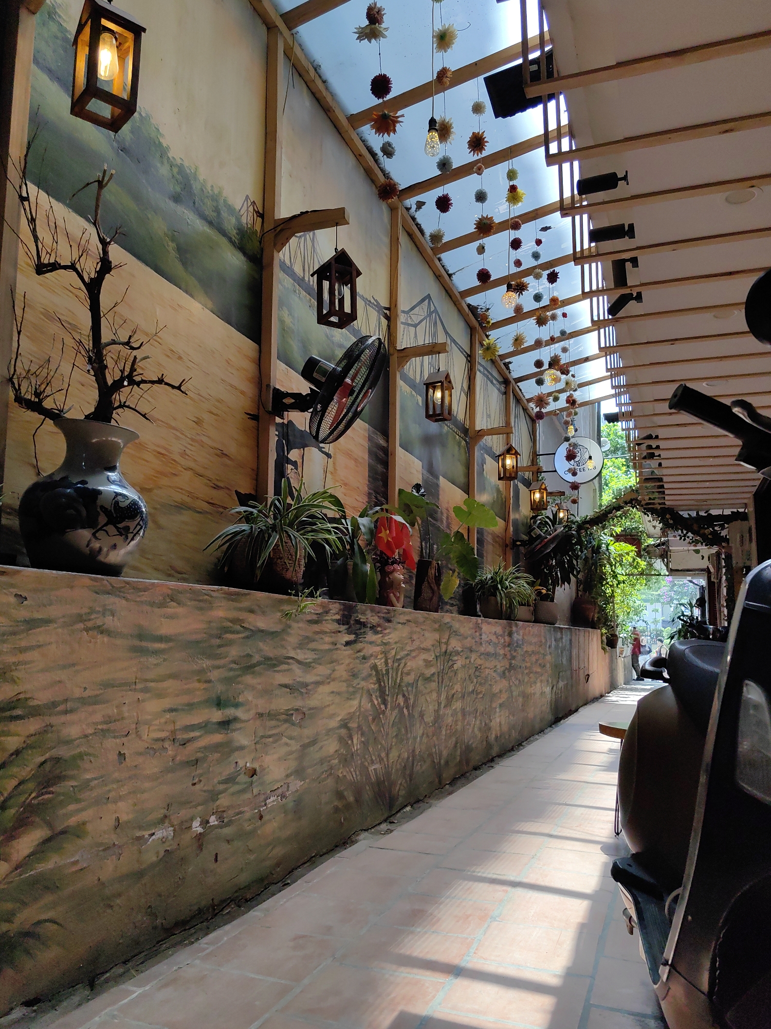 This image captures a long, narrow outdoor or semi-outdoor pathway, likely part of a cafe or restaurant in Hà Nội, Vietnam. The scene is brightly lit by natural sunlight, casting sharp shadows and suggesting a clear, sunny day, probably midday or early afternoon.
On the left, a painted wall features a mural. The upper section depicts a verdant, rolling hillside with a prominent bridge, possibly evoking a landmark like the Long Bien Bridge. The lower part of the mural is an earthy-toned landscape, showing visible signs of wear and peeling paint. A narrow ledge runs along this wall, holding various decorative items: a large, patterned vase with dried, bare tree branches, several potted green plants, and a single red flower. Rustic wooden lantern-style lights hang from wooden beams and are mounted on the wall, along with a black wall-mounted fan.
The ceiling is a notable feature, combining wooden beams with a transparent roof section that allows direct sunlight to filter through. Numerous colorful artificial flowers or pom-poms in shades of orange, yellow, red, and white, interspersed with small hanging light bulbs, adorn this transparent roof. These decorations cast intricate shadows onto the tiled pathway below.
The pathway itself is paved with light-colored square tiles. On the right side, a dark-colored scooter or motorcycle is parked, its seat and handlebars visible in the foreground, partially obscuring the view further down the corridor. As the path recedes, it narrows and is flanked by more potted plants and hanging vines, creating a lush, green archway. A circular white sign is visible, displaying the text "COFFEE 1.8" along with an illustrative logo. In the far distance, beyond the path's end, blurred figures, including one person in a red top, can be seen, suggesting an outdoor area with activity. The overall ambiance is artistic, natural, and inviting.