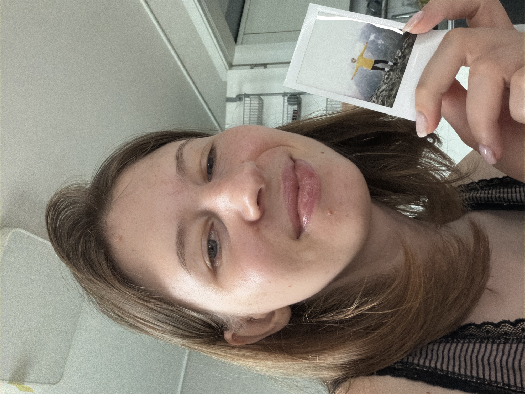 A young woman with shoulder-length brown hair is taking a selfie indoors. She is holding a small polaroid photograph in her right hand, showing a person in a yellow jacket standing on a mountain. The woman is looking towards the camera with a slight smile. Her clothing appears to be a black lace-trimmed top. The background suggests an indoor setting, possibly a room with shelves and a doorway visible. The overall scene is brightly lit. The location is specified as Dongducheon-si, South Korea.