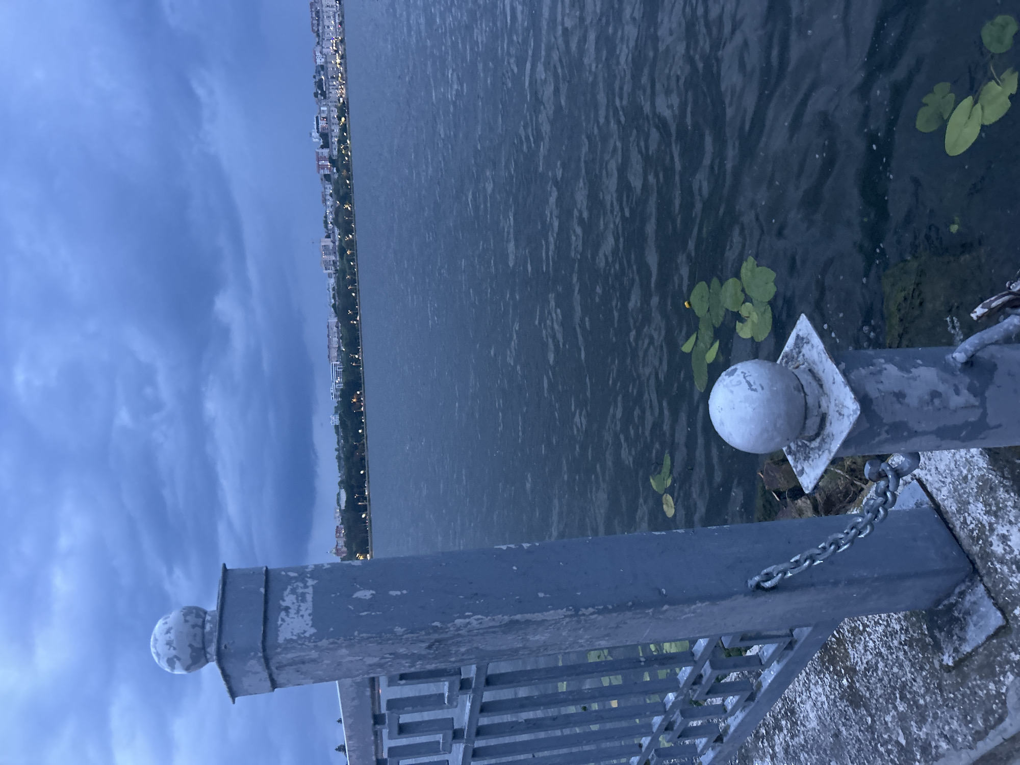 The image depicts a twilight scene at the Ternopil Lake in Ternopil, Ukraine. In the foreground, a weathered gray metal fence with decorative geometric patterns runs along a concrete walkway. A substantial gray metal post supporting the fence is secured with a chain. The post is topped with a weathered spherical finial. On the left side of the image, the fence extends into the foreground. The lake stretches out in the midground, its surface rippled by a gentle breeze. Lily pads, some with small yellow flowers, float on the water. In the background, the Ternopil cityscape is visible, with buildings of various sizes lining the shore, illuminated by streetlights and interior lights, indicating it is dusk or early evening. The sky above is overcast with dark, heavy clouds, contributing to the somber atmosphere of the scene. No people are visible in the image.