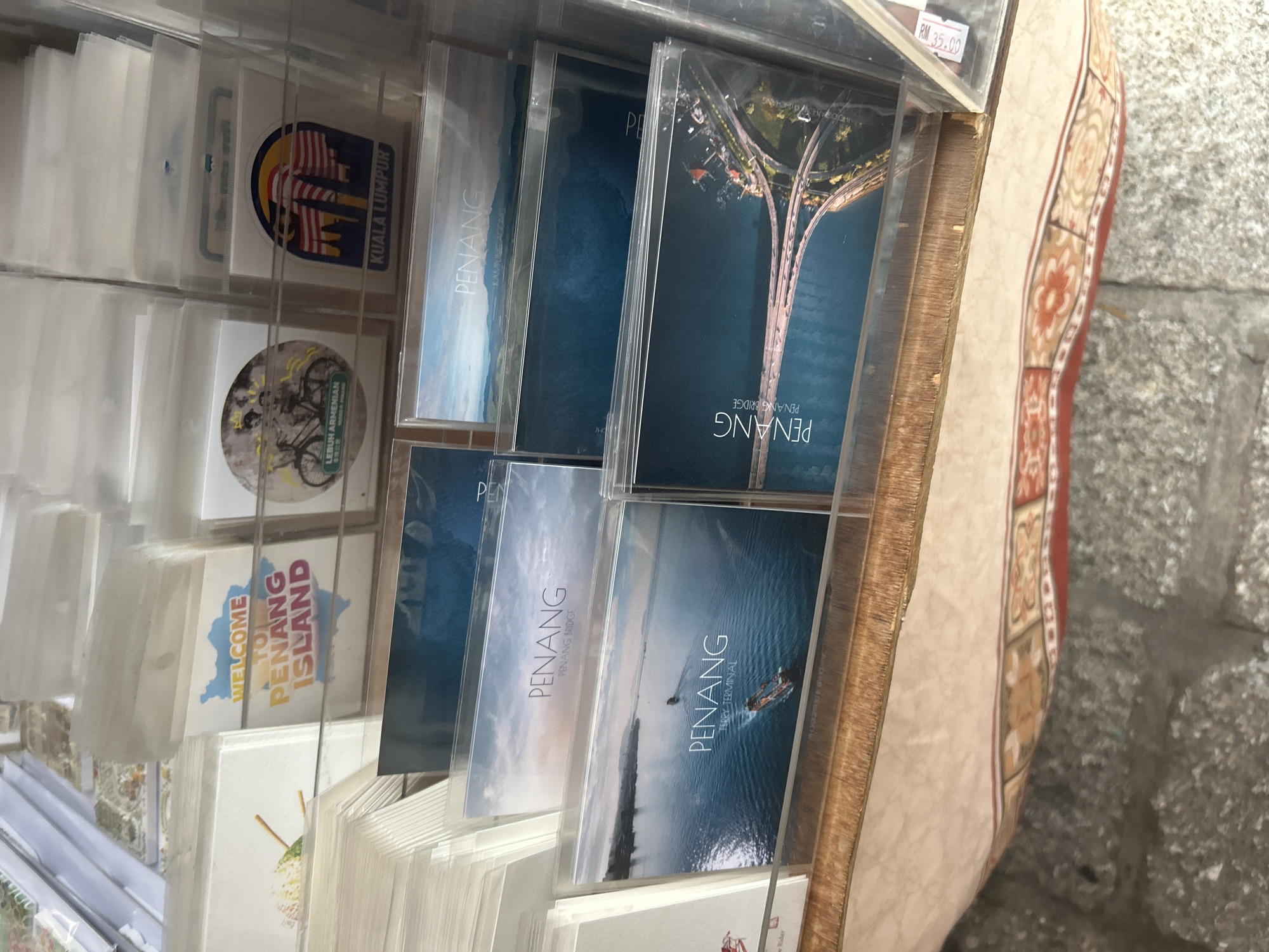 The image shows a display of souvenirs, likely postcards and small prints, for sale in what appears to be a shop or market stall in George Town, Malaysia. The items are arranged in clear plastic sleeves and stacked in transparent display racks. Prominently featured are various postcards depicting scenes of Penang Island, including the Penang Bridge and the Penang Ferry Terminal, with the text "PENANG" clearly visible on several of them. One postcard has the text "WELCOME TO PENANG ISLAND" overlaid on a map of Penang. Another item shows a sticker or postcard with the Malaysian flag and the text "KUALA LUMPUR". The background suggests an indoor setting with further merchandise stacked behind the main display. The overall presentation is commercial, aimed at tourists.