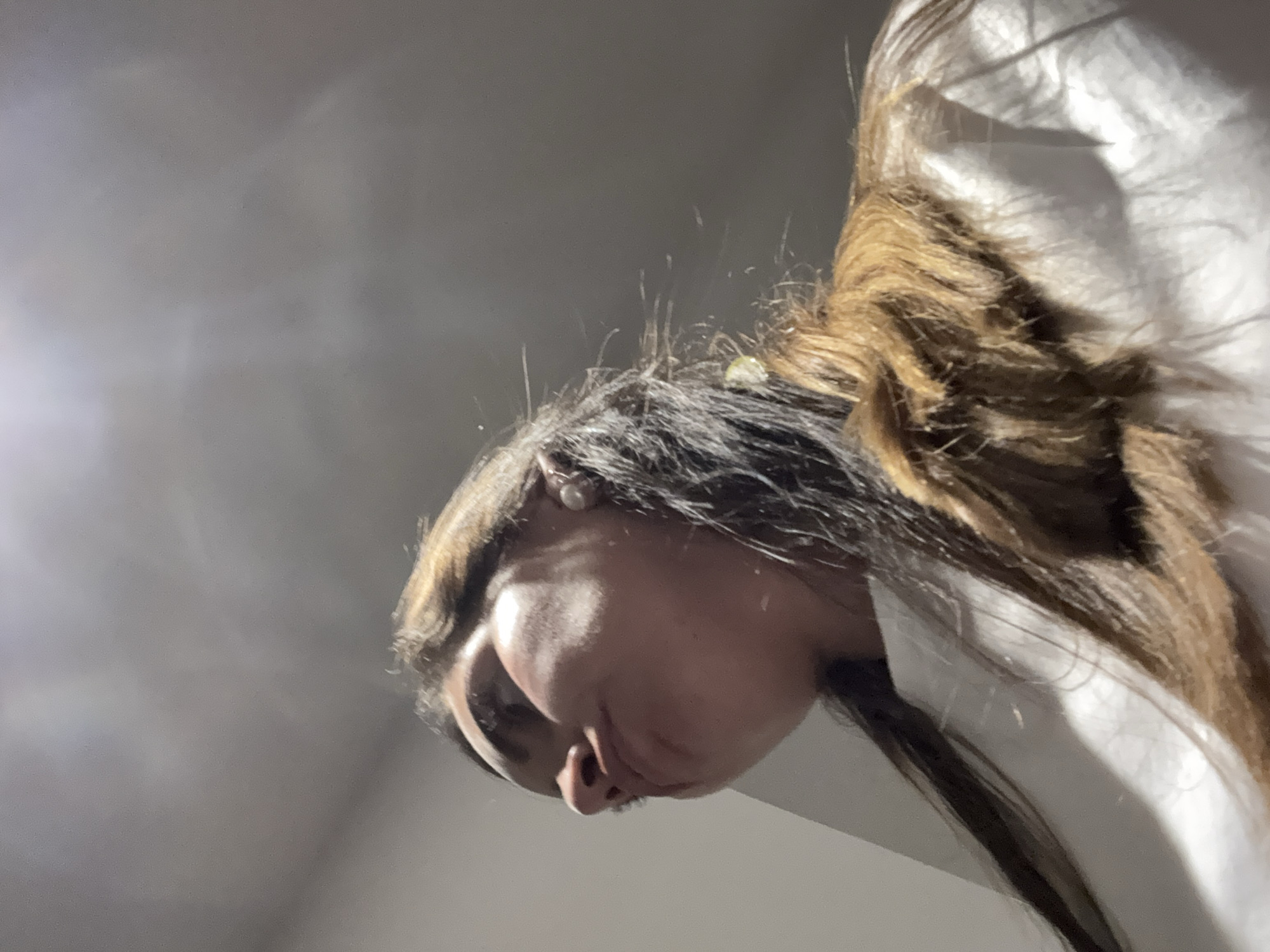 The image shows a person from a low-angle perspective. The subject has brown and black hair and is wearing a white shirt. The background is a blurred grey. There is no information in the image that suggests it was taken in San Bernardino Tlaxcalancingo, Mexico.
