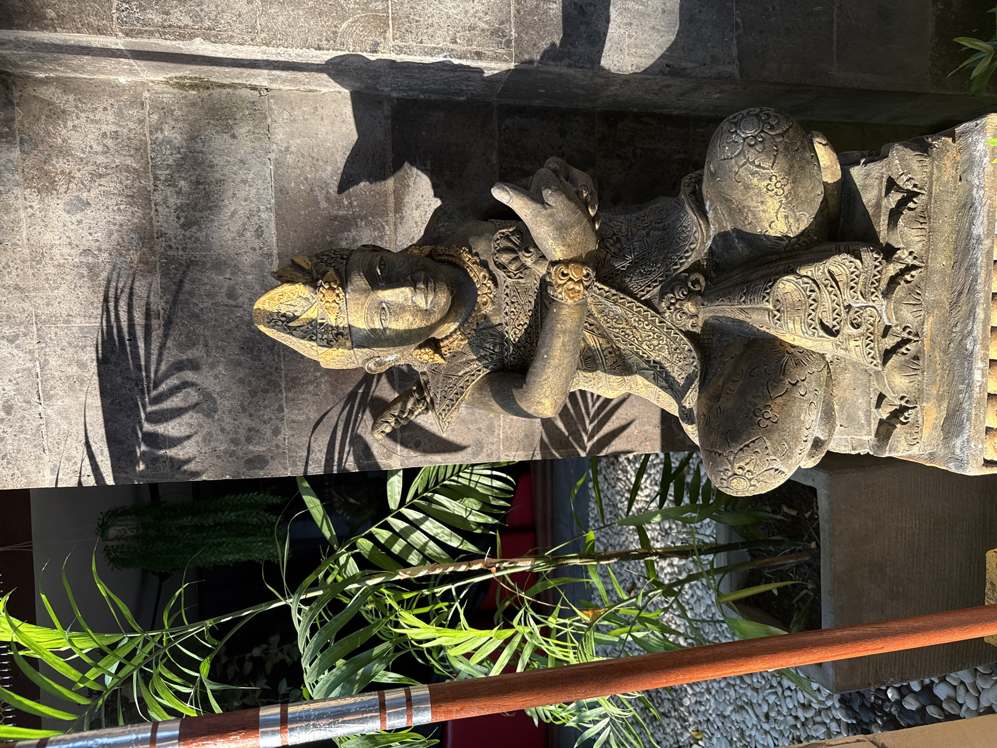 A stone statue of a seated figure wearing traditional Balinese attire with golden accents is positioned in front of a textured, grey stone wall. The statue is detailed, with intricate patterns on its clothing, a headdress, and jewelry. To the left of the statue, a tall potted plant with vibrant green fronds receives direct sunlight, casting shadows on the wall. A wooden pole with metallic rings stands vertically in the foreground, partially obscuring the plant. The ground around the pot is covered with small, smooth white stones. The lighting suggests it is daytime, with strong shadows indicating a low sun angle. The overall scene depicts an outdoor setting, likely a garden or courtyard area within a resort or private residence in Kuta Selatan, Indonesia, given the architectural style and foliage.